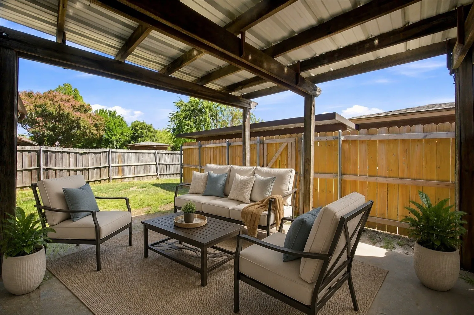 Covered patio with a corrugated metal roof, wood beam supports, concrete flooring, and a privacy fence