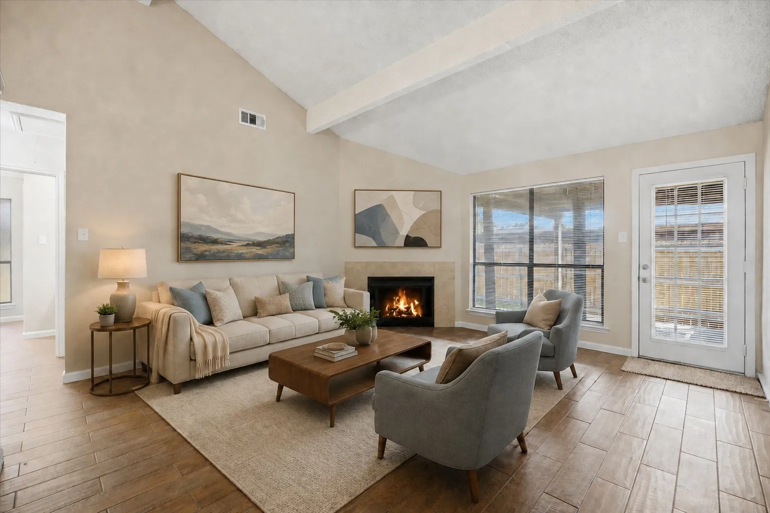Vaulted ceiling with exposed beams, wood-finish flooring, corner fireplace with stone surround, large window, and a glass-paneled door