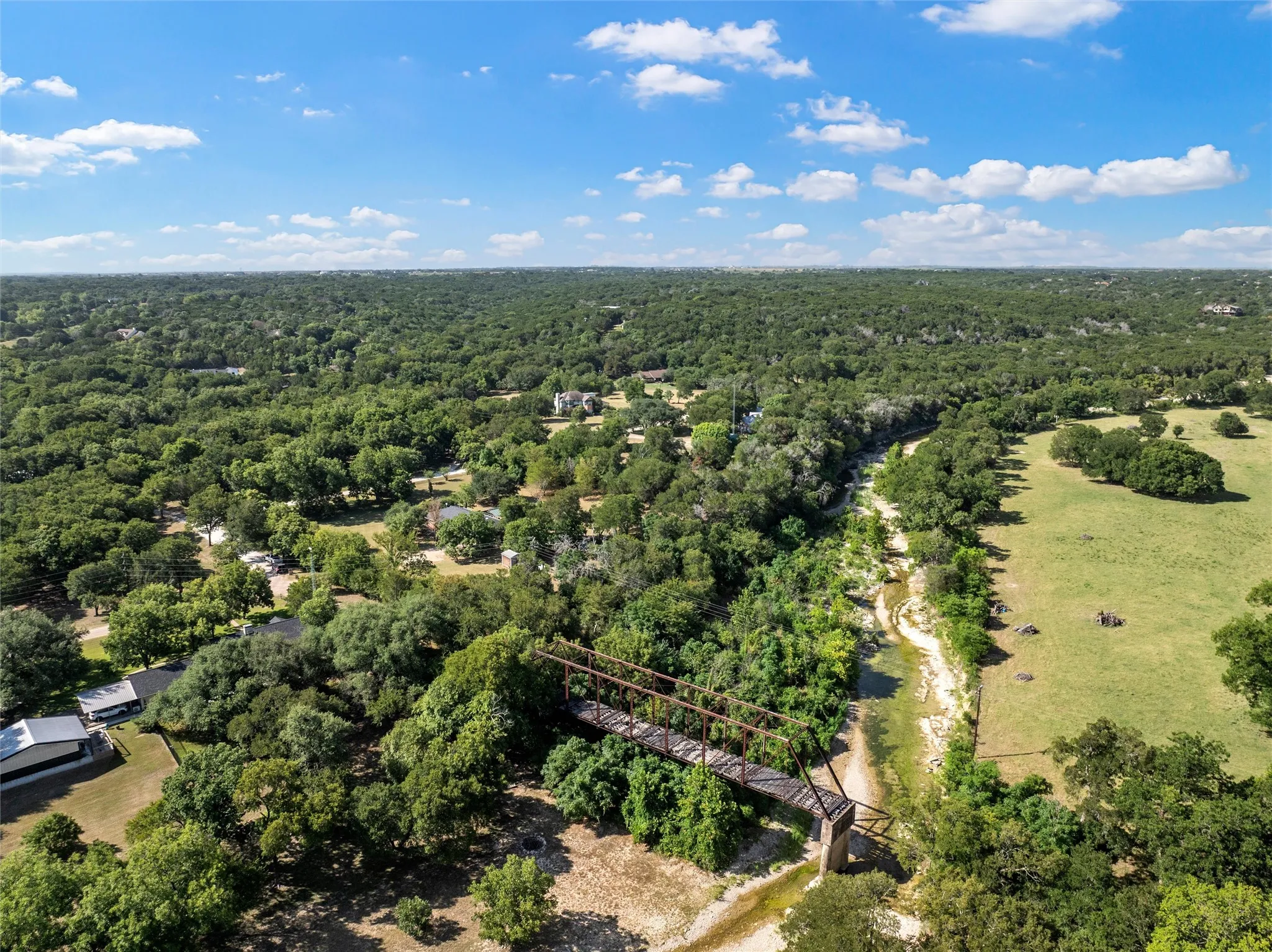 Aerial landscape featuring extensive tree coverage, a natural waterway, a truss bridge, and open grassy areas