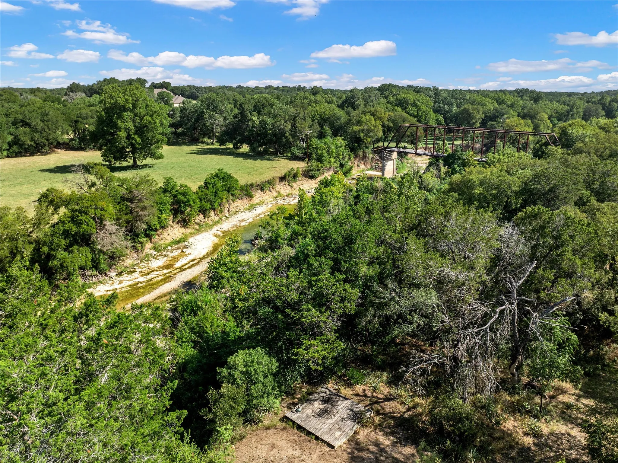 Expansive natural landscape featuring a meandering creek, mature trees, and a distant metal truss bridge