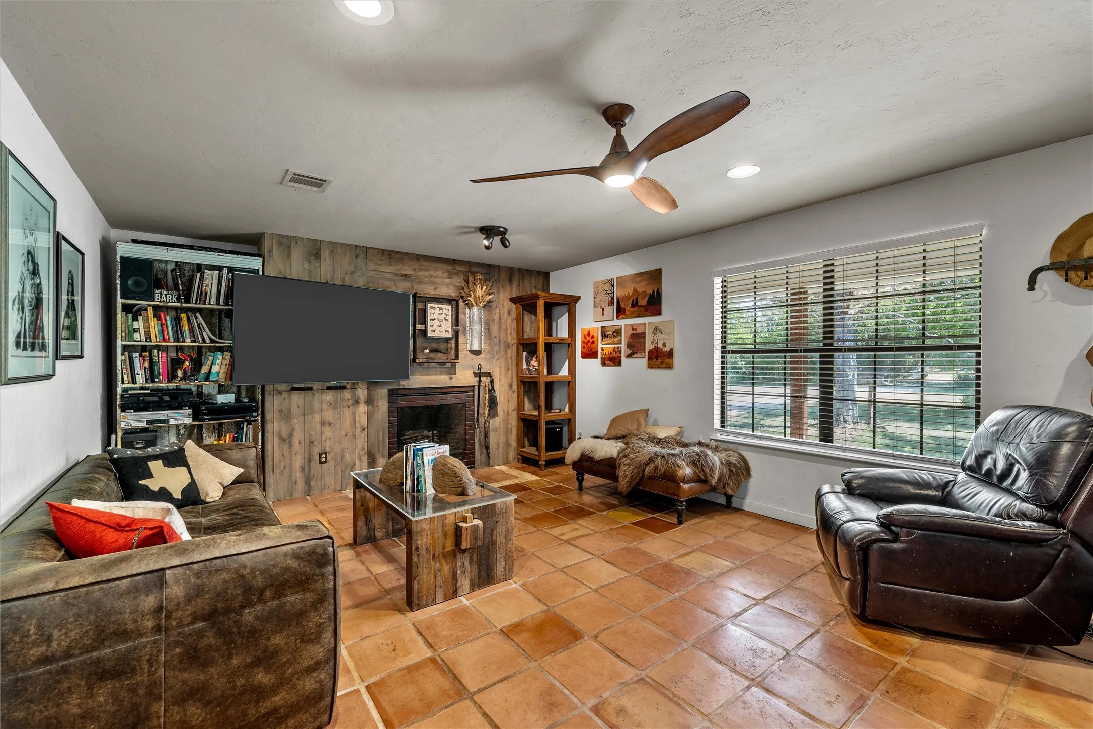 Terracotta tile flooring, wood-paneled accent wall, built-in fireplace, and a ceiling fan with integrated lighting