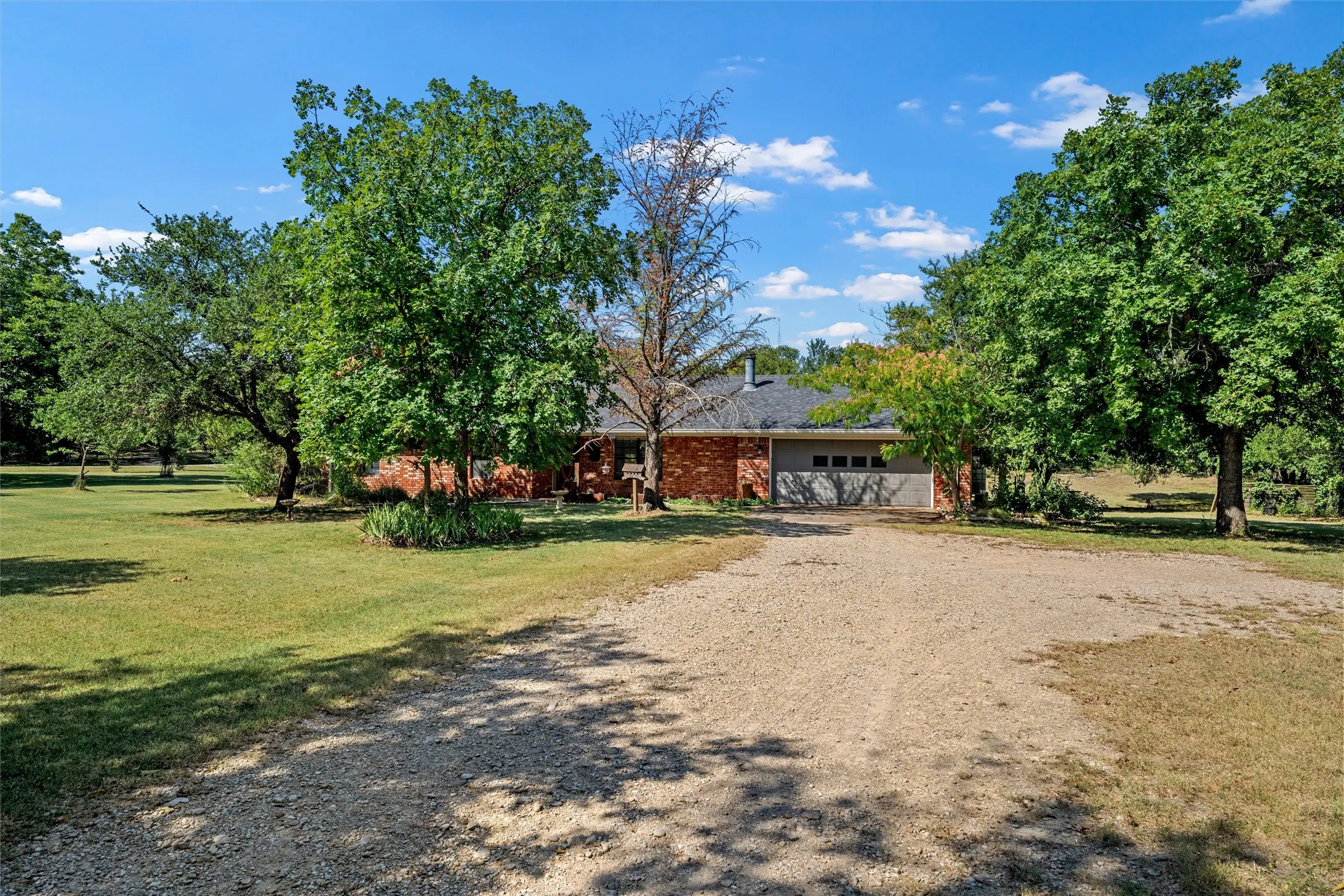 Brick exterior with a gray-sided garage, gravel driveway, and mature landscaping