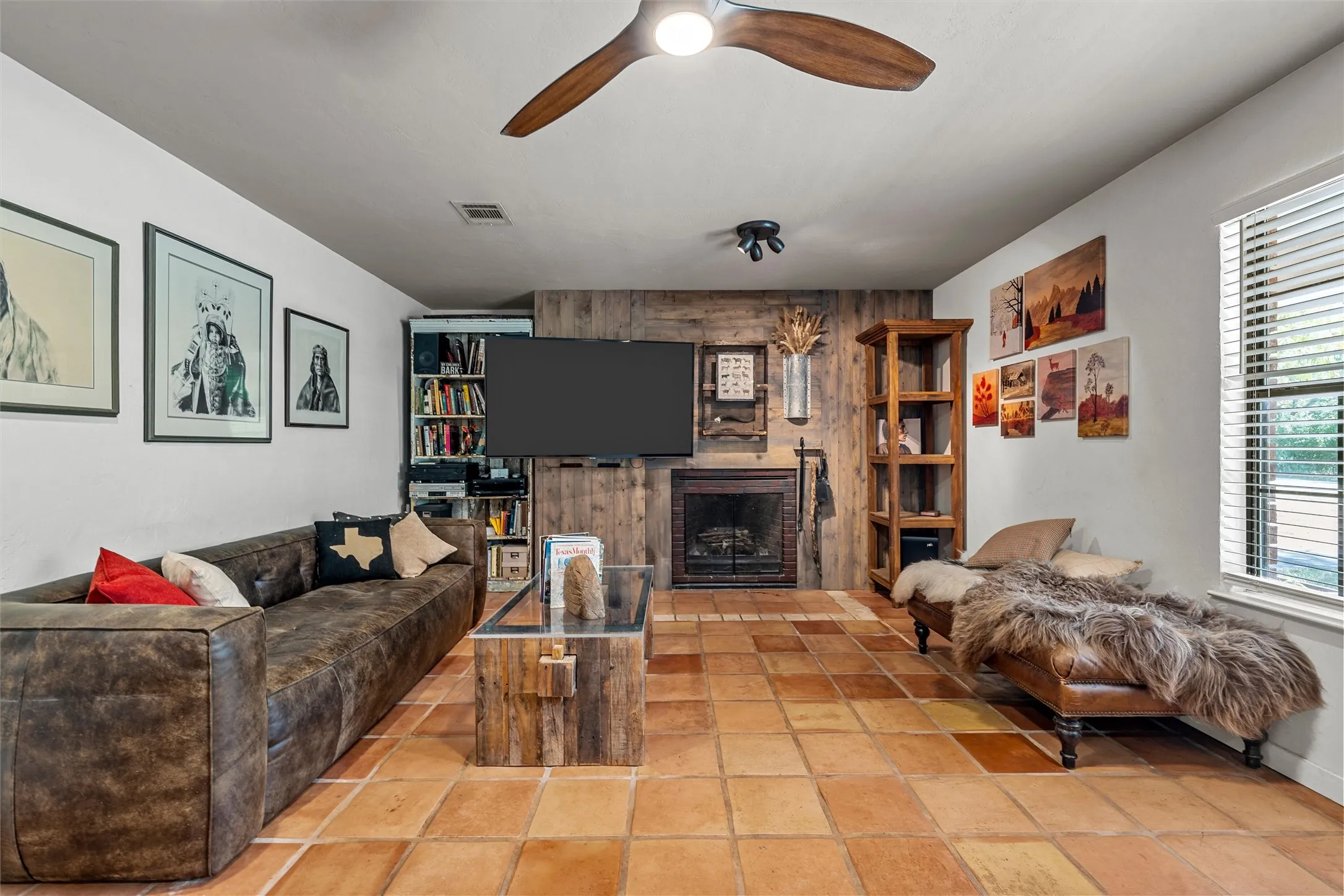 Terracotta tile flooring, wood-paneled accent wall, built-in fireplace, and a ceiling fan with integrated lighting
