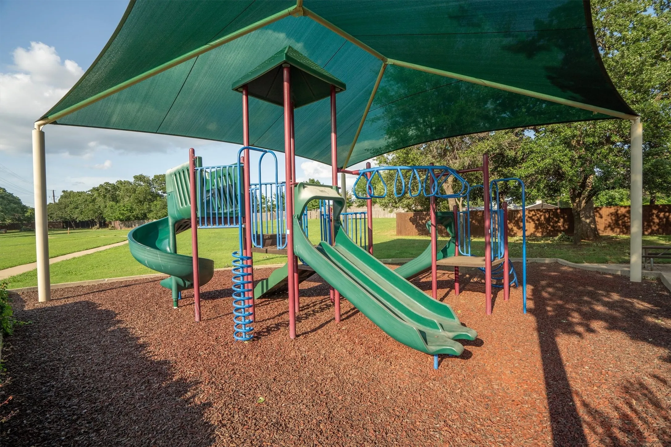 Playground area featuring a shade structure, multiple slides, climbing elements, and a ground cover of reddish-brown material
