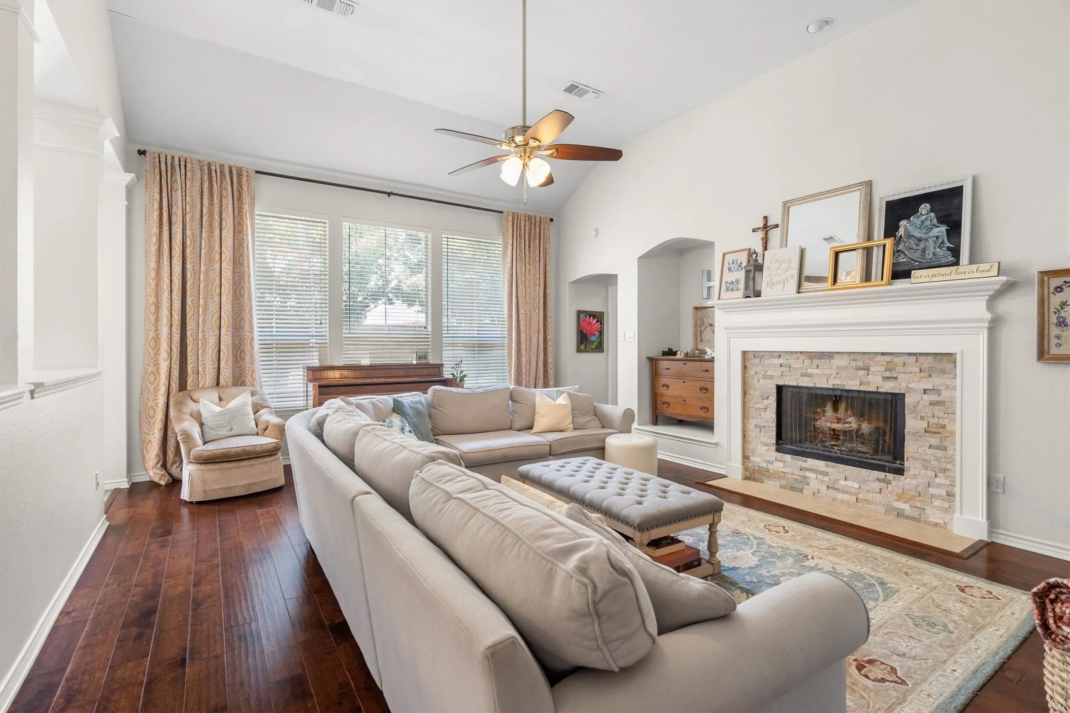 Vaulted ceiling with a ceiling fan, wood-finish flooring, a fireplace with a stone surround and white mantel, and multiple windows