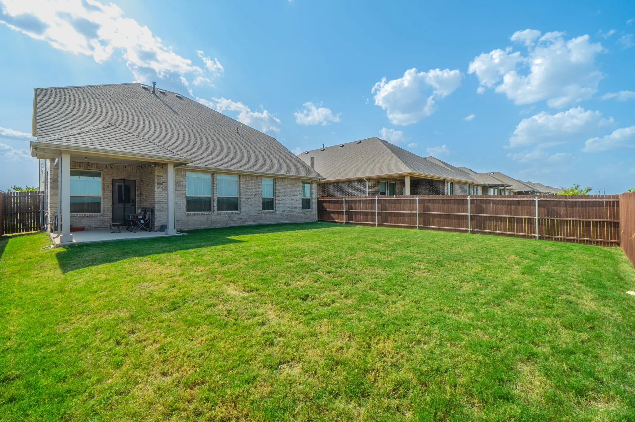 Expansive lawn area with a wood privacy fence, brick exterior, covered patio, and multiple windows