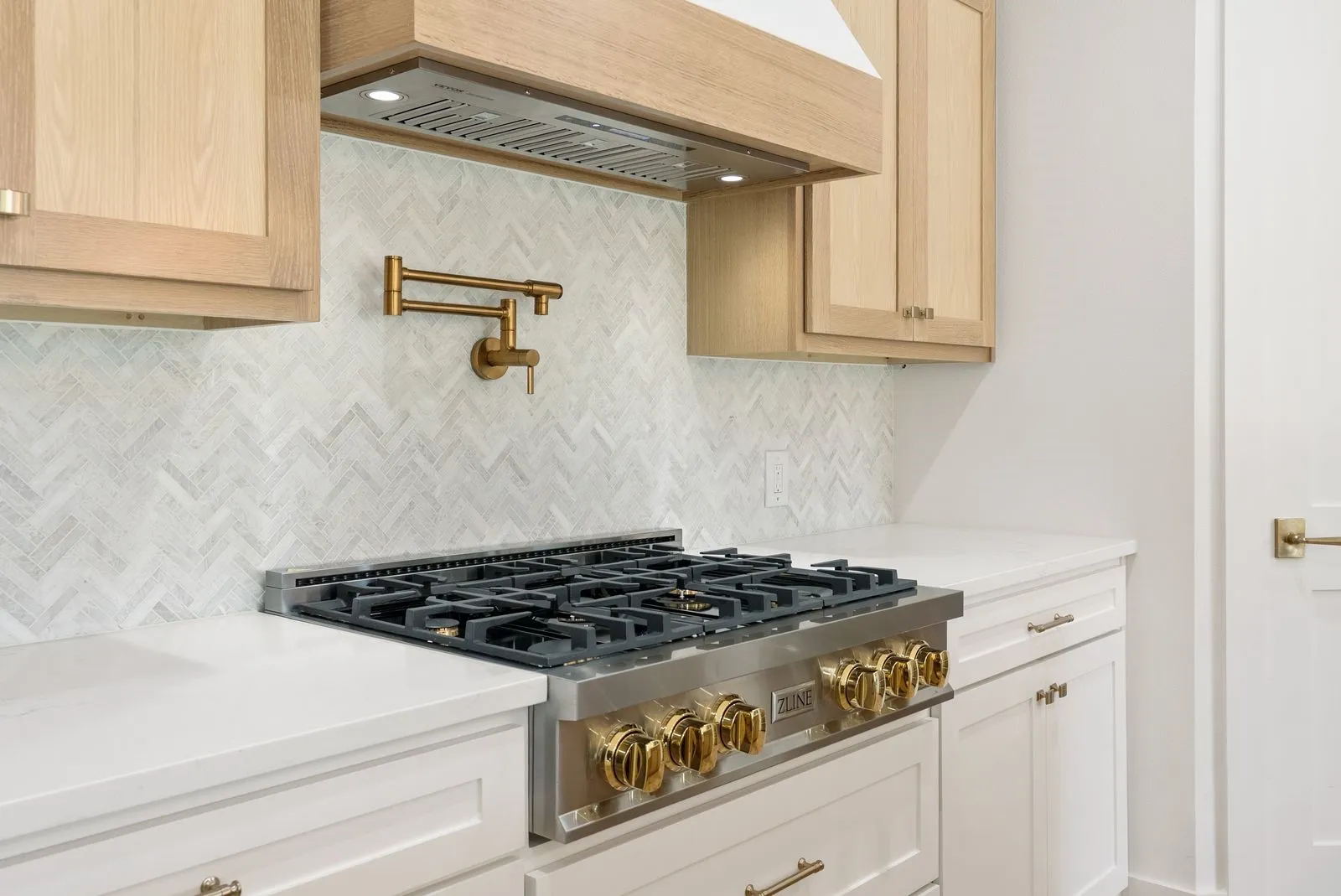 Gourmet kitchen featuring a professional-grade gas range, herringbone tile backsplash, gold-tone pot filler, light wood-finish cabinetry, and white countertops
