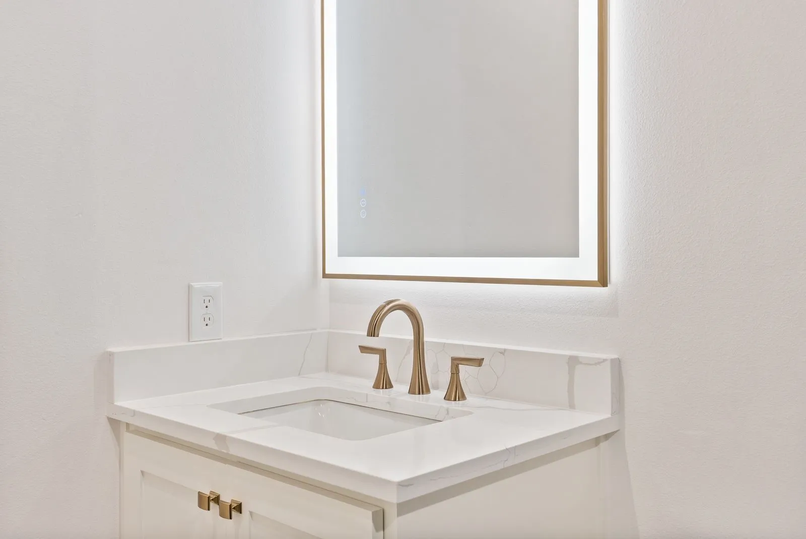 Vanity featuring an integrated sink, light-colored countertop with subtle veining, brushed gold-tone faucet, and a back-lit mirror