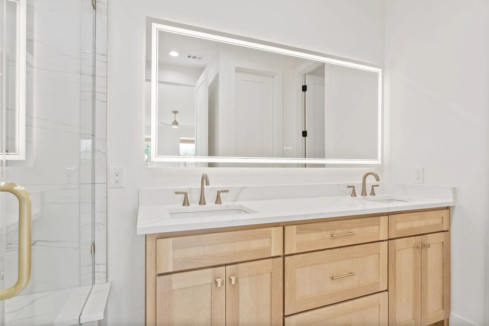 Dual vanity with a white countertop and natural wood-finish cabinetry, illuminated mirror, and a clear glass shower enclosure featuring a built-in bench