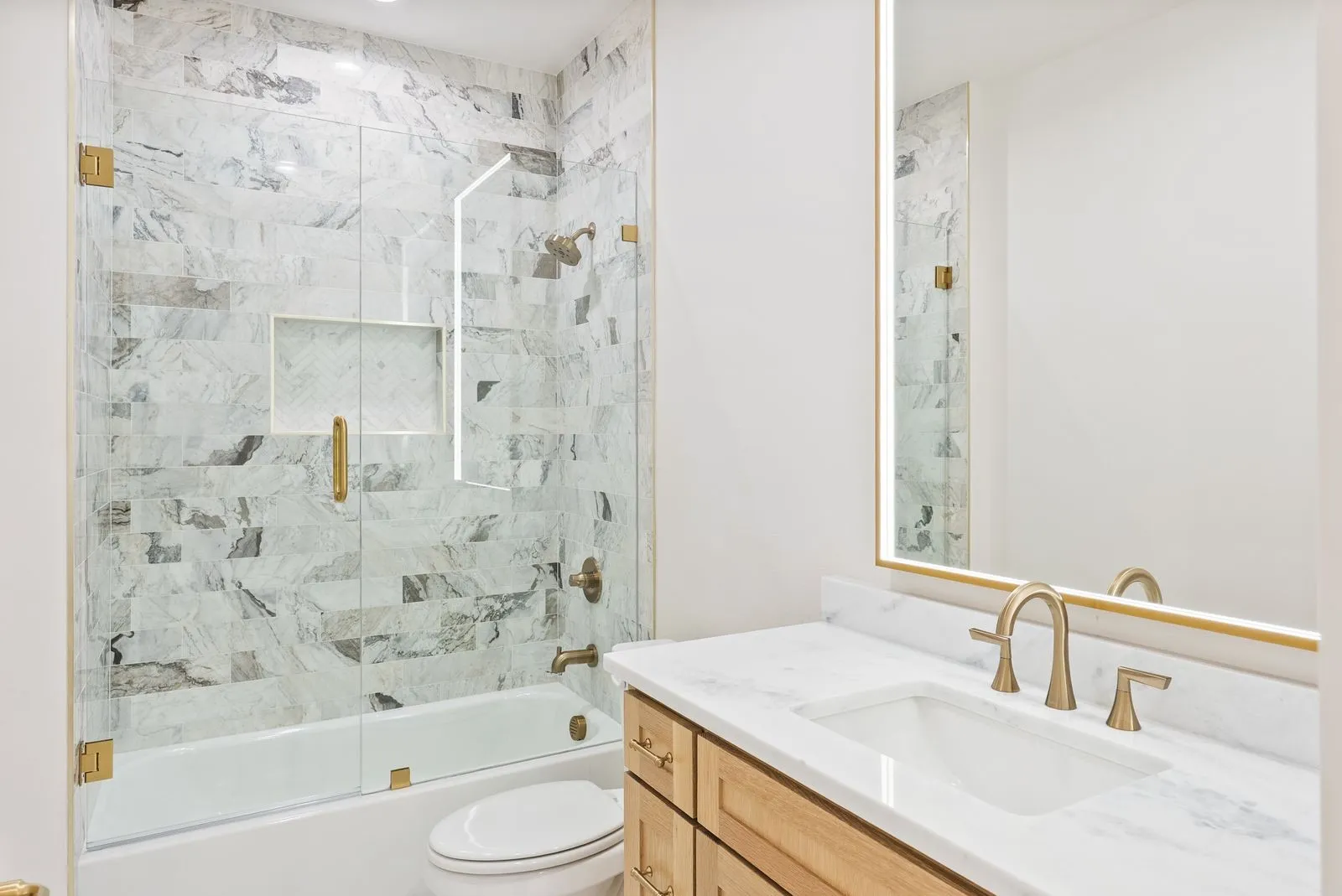 Bathtub and shower combination with glass enclosure, marble-look tiled walls, and a recessed niche