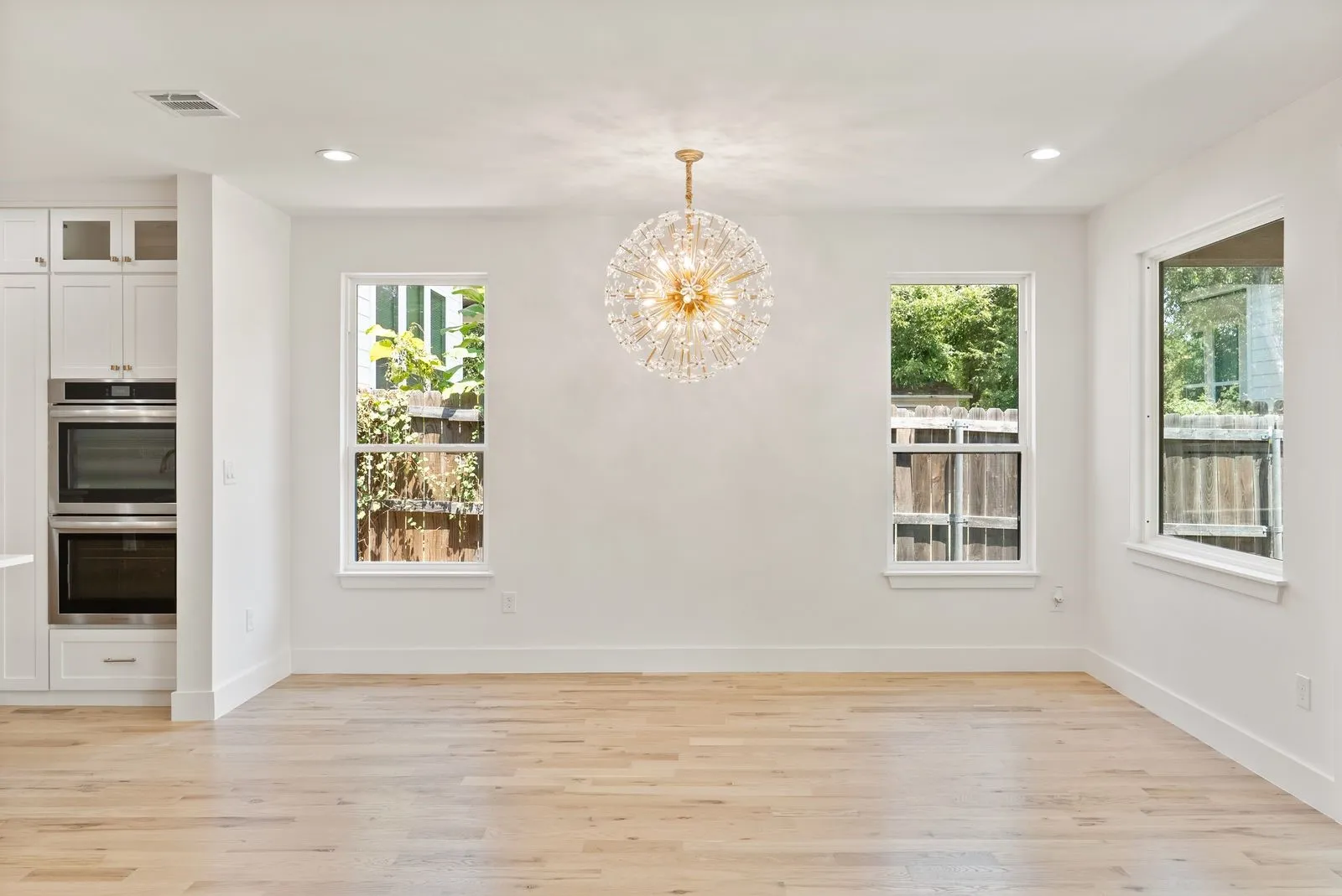 Light wood-finish flooring, recessed ceiling lighting, a crystal chandelier, built-in double ovens, and white cabinetry