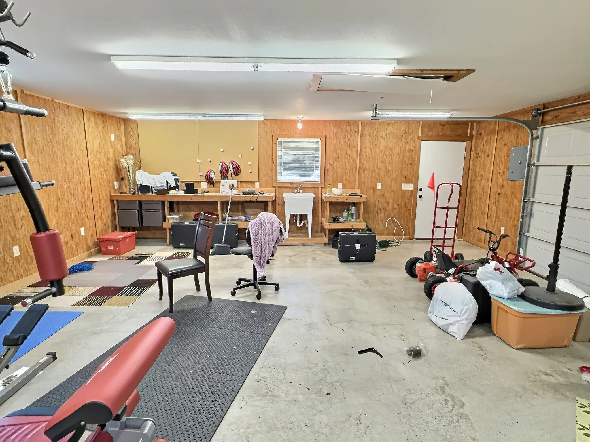 Spacious shop featuring light wood paneling, concrete flooring, ample overhead lighting, a utility sink, and a built-in workbench