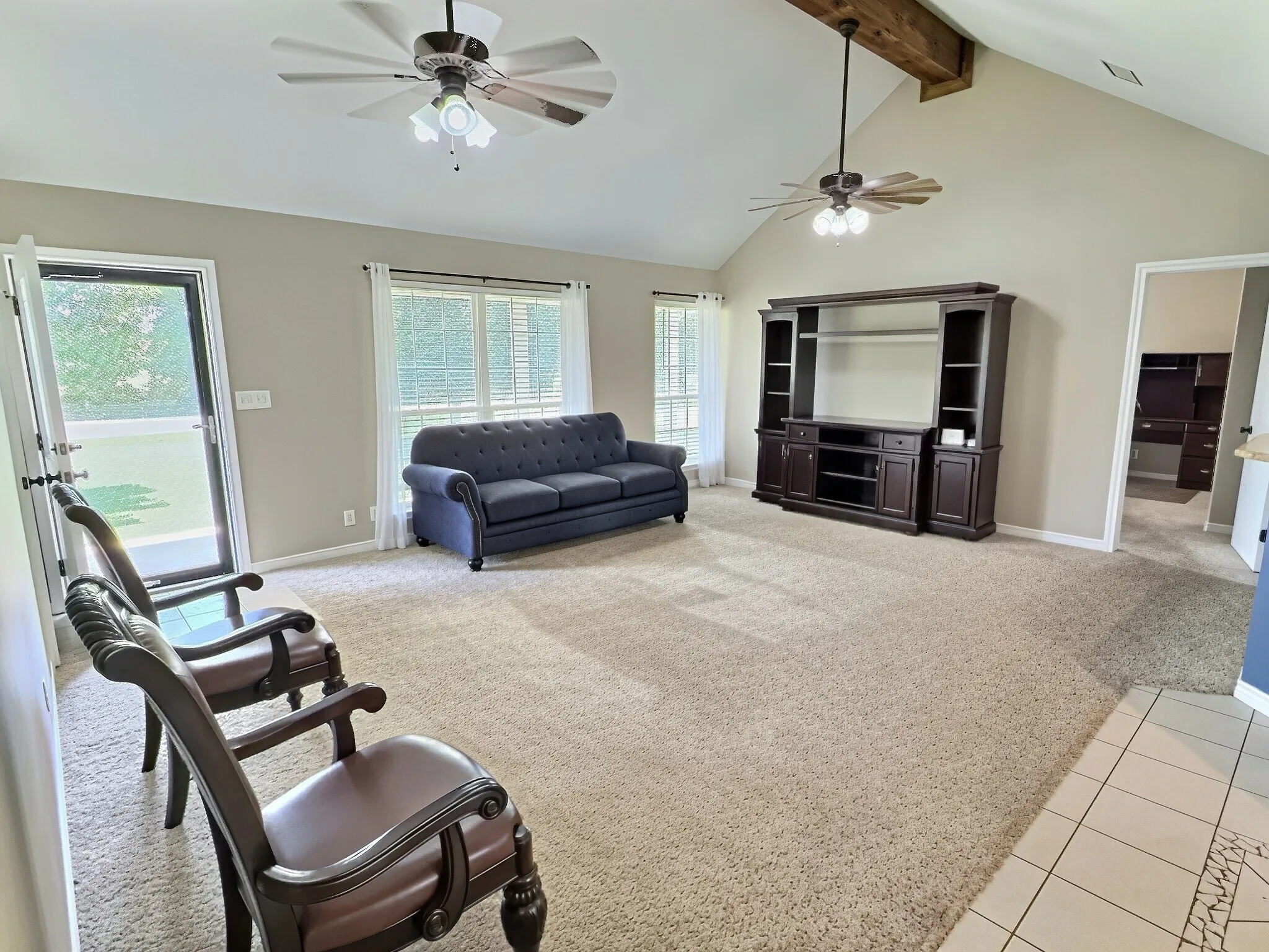 Vaulted ceilings with exposed wood beam, two ceiling fans with lighting, large windows with blinds, and light-toned carpeting