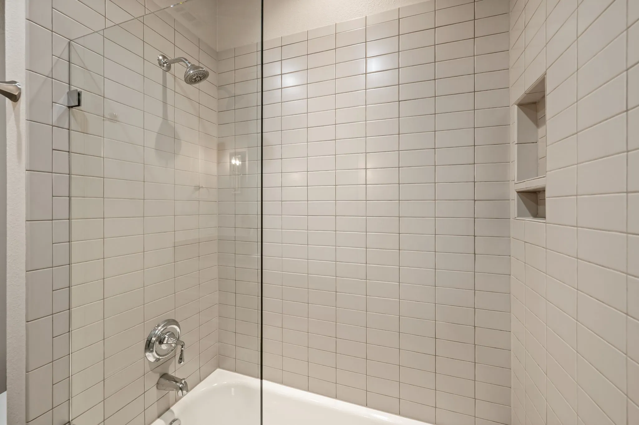 Shower-over-tub combination with light-toned tiled walls, built-in shower niches, and a frameless glass panel