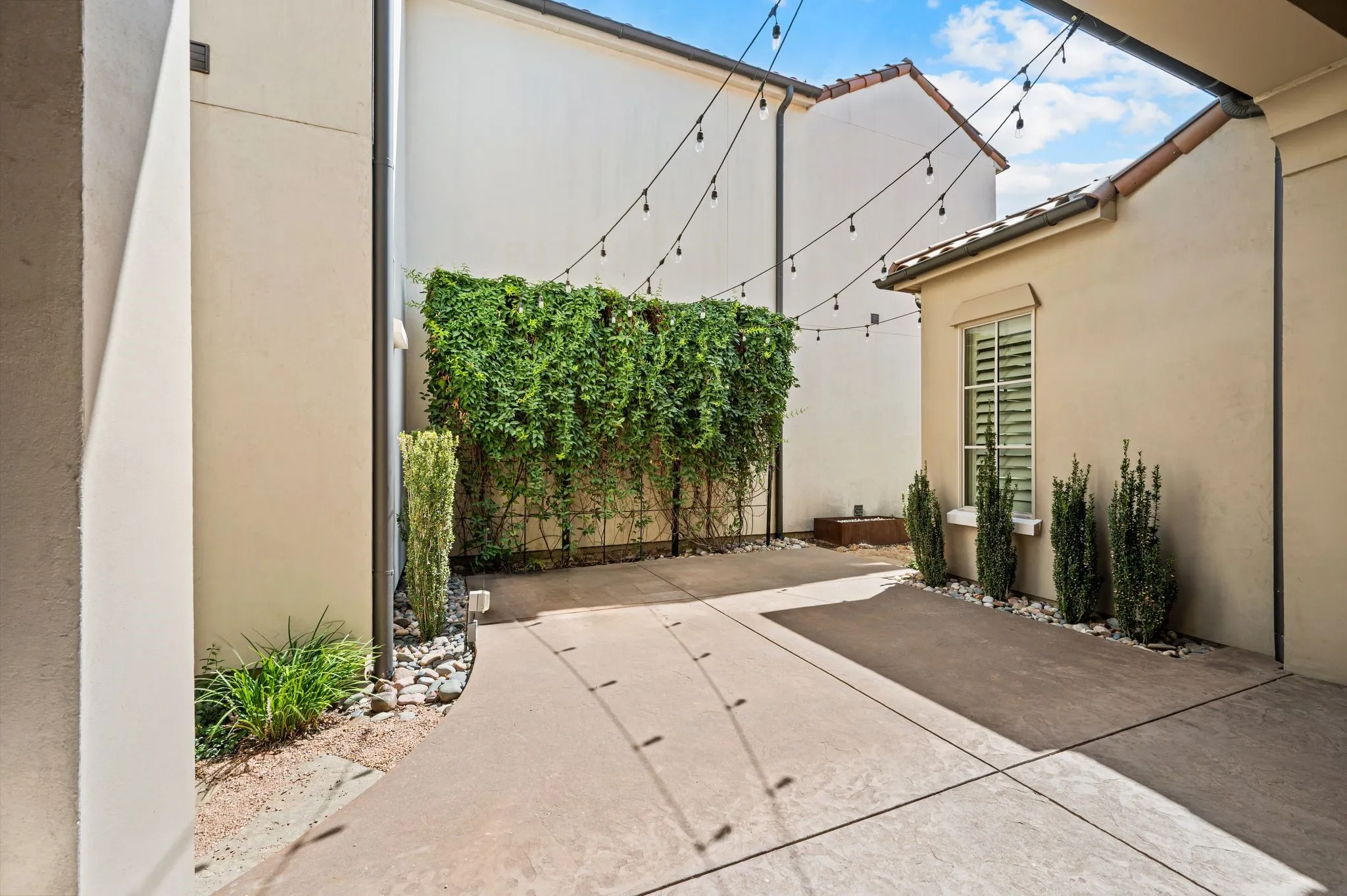 Modern courtyard featuring a retractable shade structure with integrated string lighting, composite decking, drought-tolerant landscaping with cascading ivy wall feature, and columnar evergreen plantings
