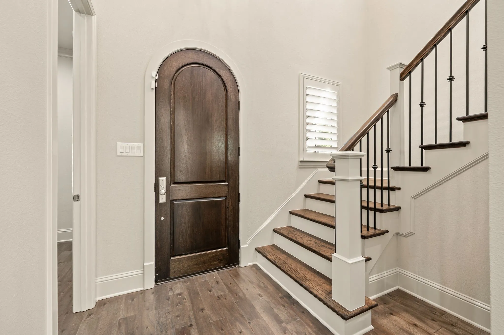 Arched wood-panel door, wood-finish flooring, staircase with wood treads, black metal balusters, and a window with plantation shutters