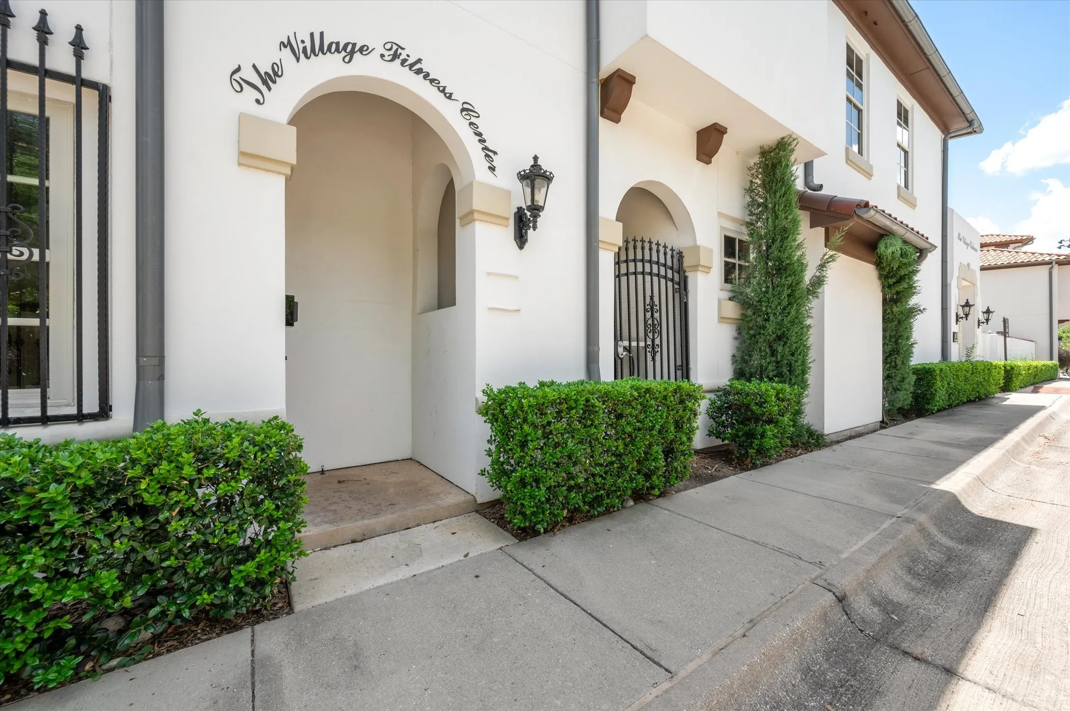 Entrance to Community Fitness Center featuring a graceful arched entryway with decorative tile detailing, white stucco exterior, black wrought-iron accents, manicured boxwood hedging, and professional hardscape with clean concrete walkway
