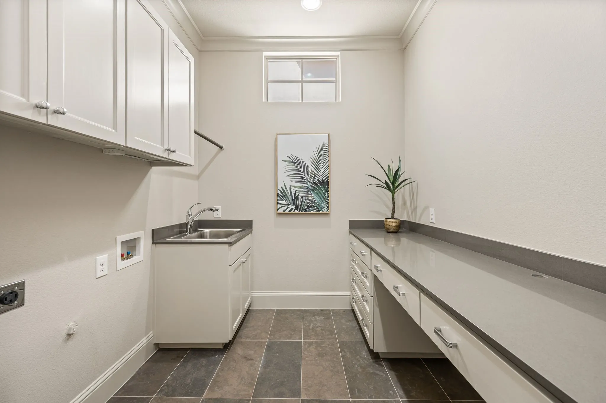 Laundry area featuring stone-look tile flooring, extensive countertop space with integrated sink, white cabinetry with chrome hardware, and a high window