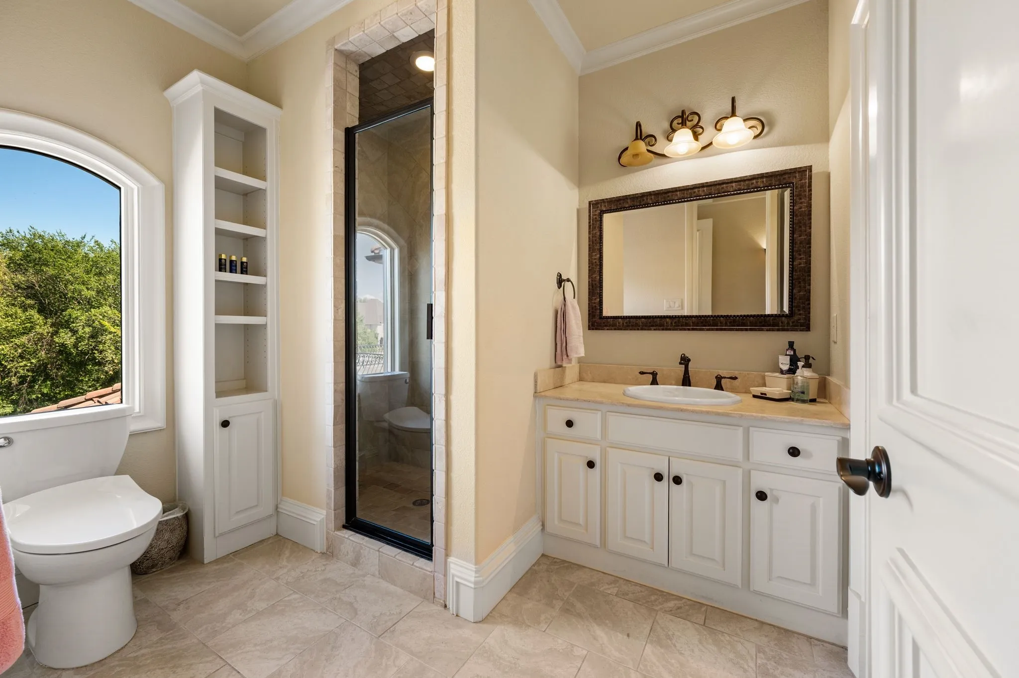 White vanity with an integrated sink and light-toned countertop, framed mirror with sconce lighting, glass-enclosed shower, white built-in storage cabinet, and arched window