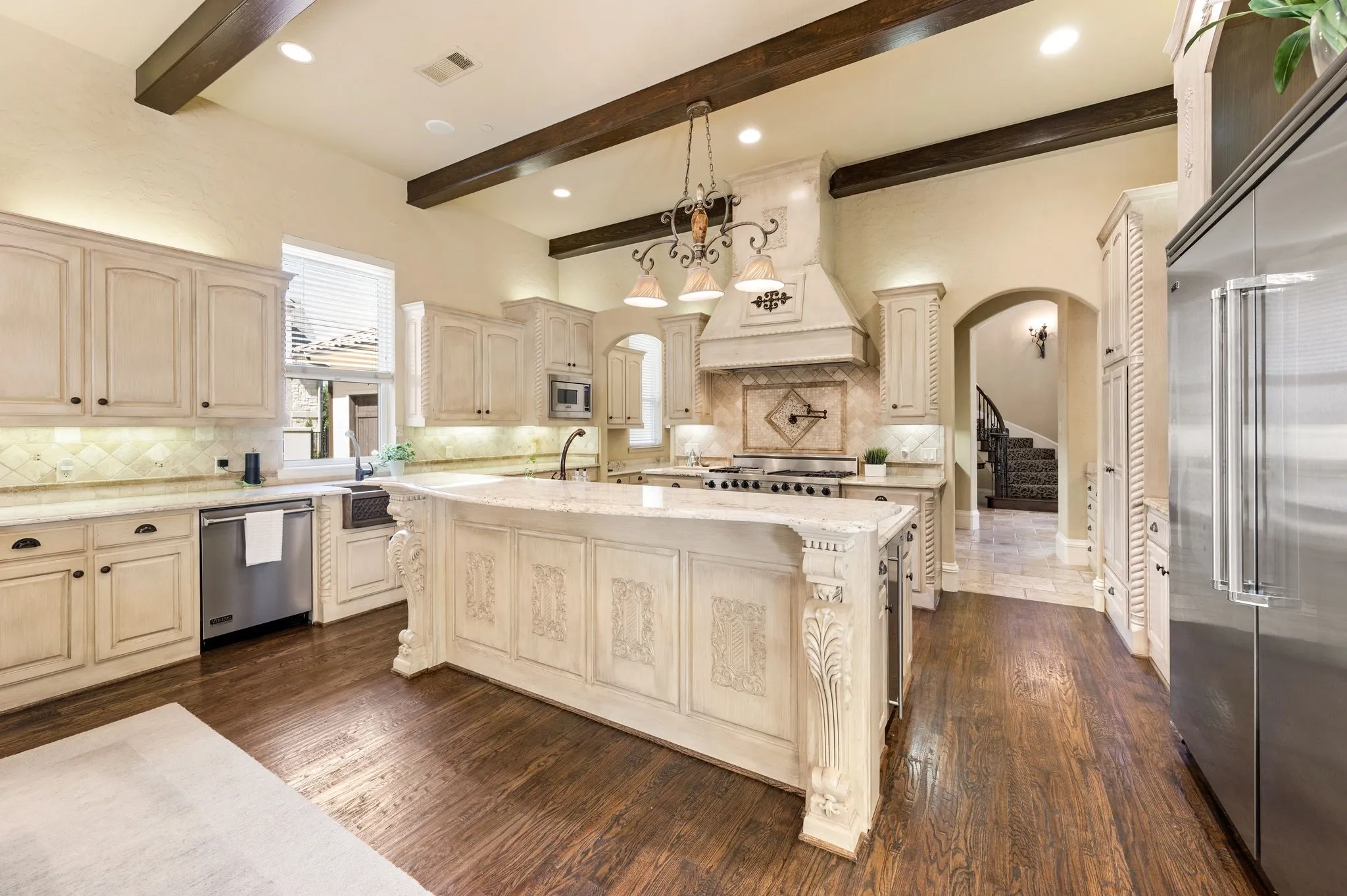 Kitchen featuring dark wood-finish flooring, vaulted ceiling with exposed beams, light cabinetry, stainless steel appliances, and a large center island with ornate detailing