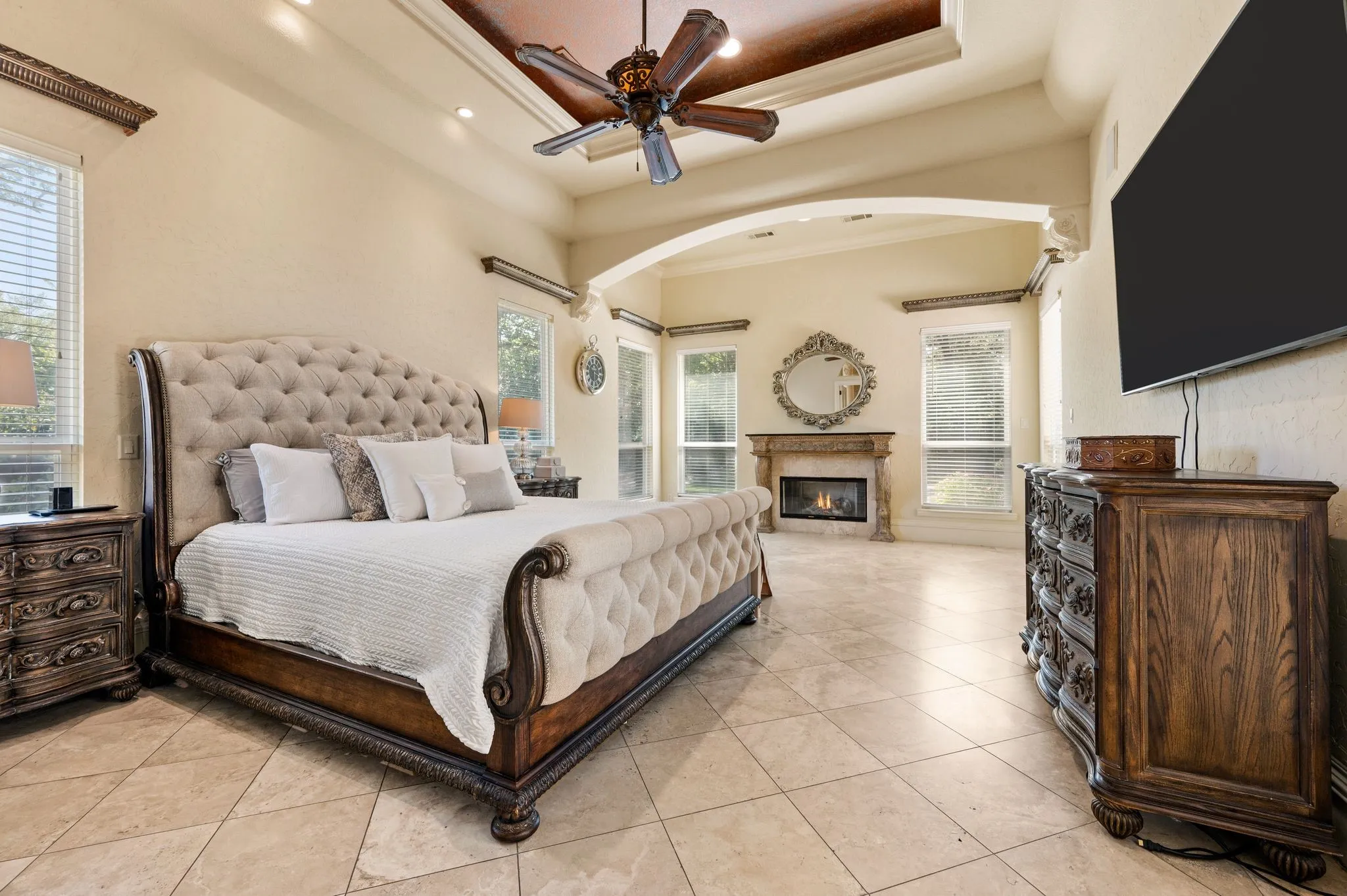 Tray ceiling with recessed lighting and a ceiling fan, arched entry, tiled flooring, and a fireplace with a stone surround