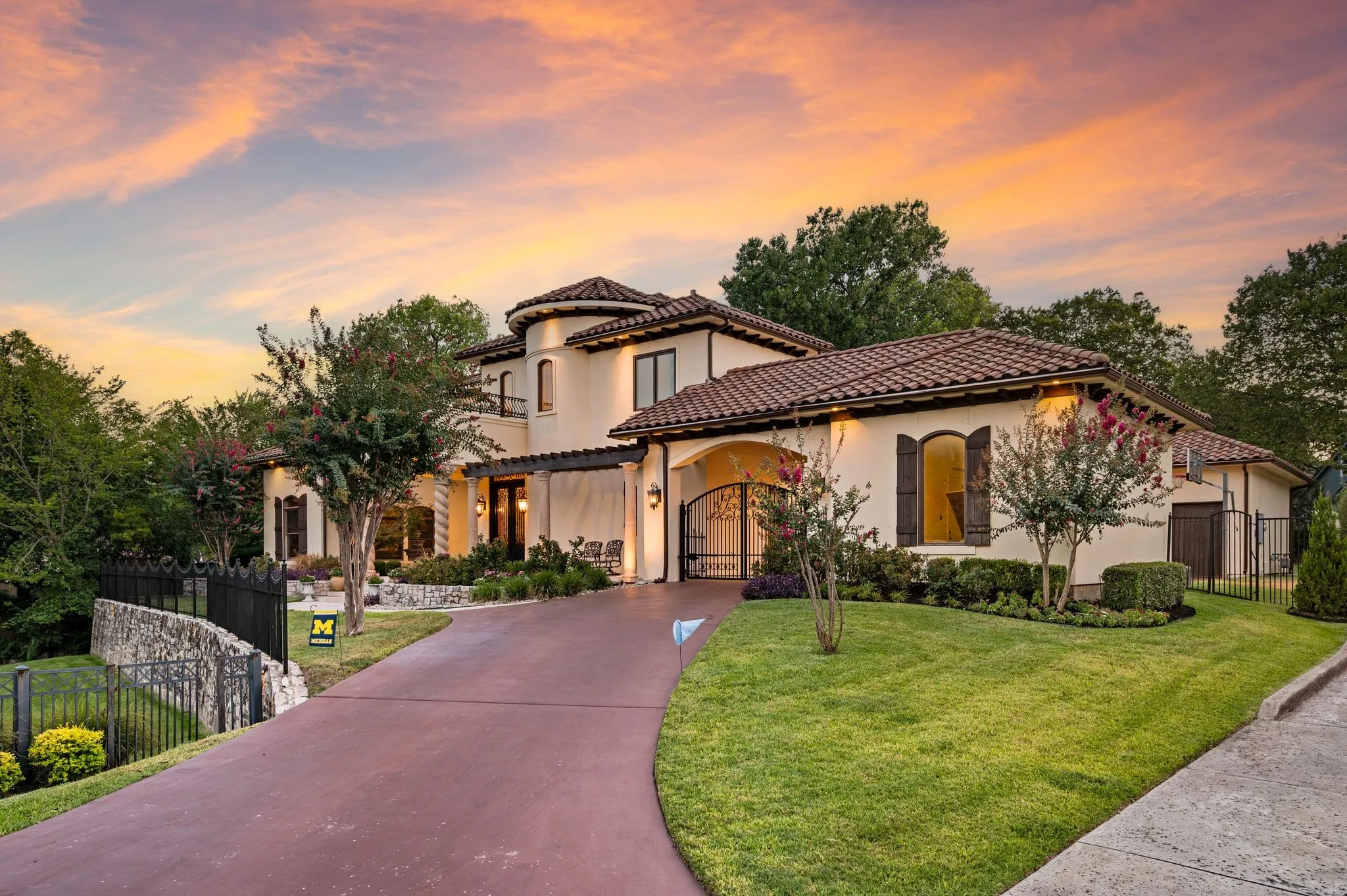 Mediterranean-style architecture with a terracotta tile roof, stucco exterior, ornate iron entry gates, a paved driveway, and mature landscaping