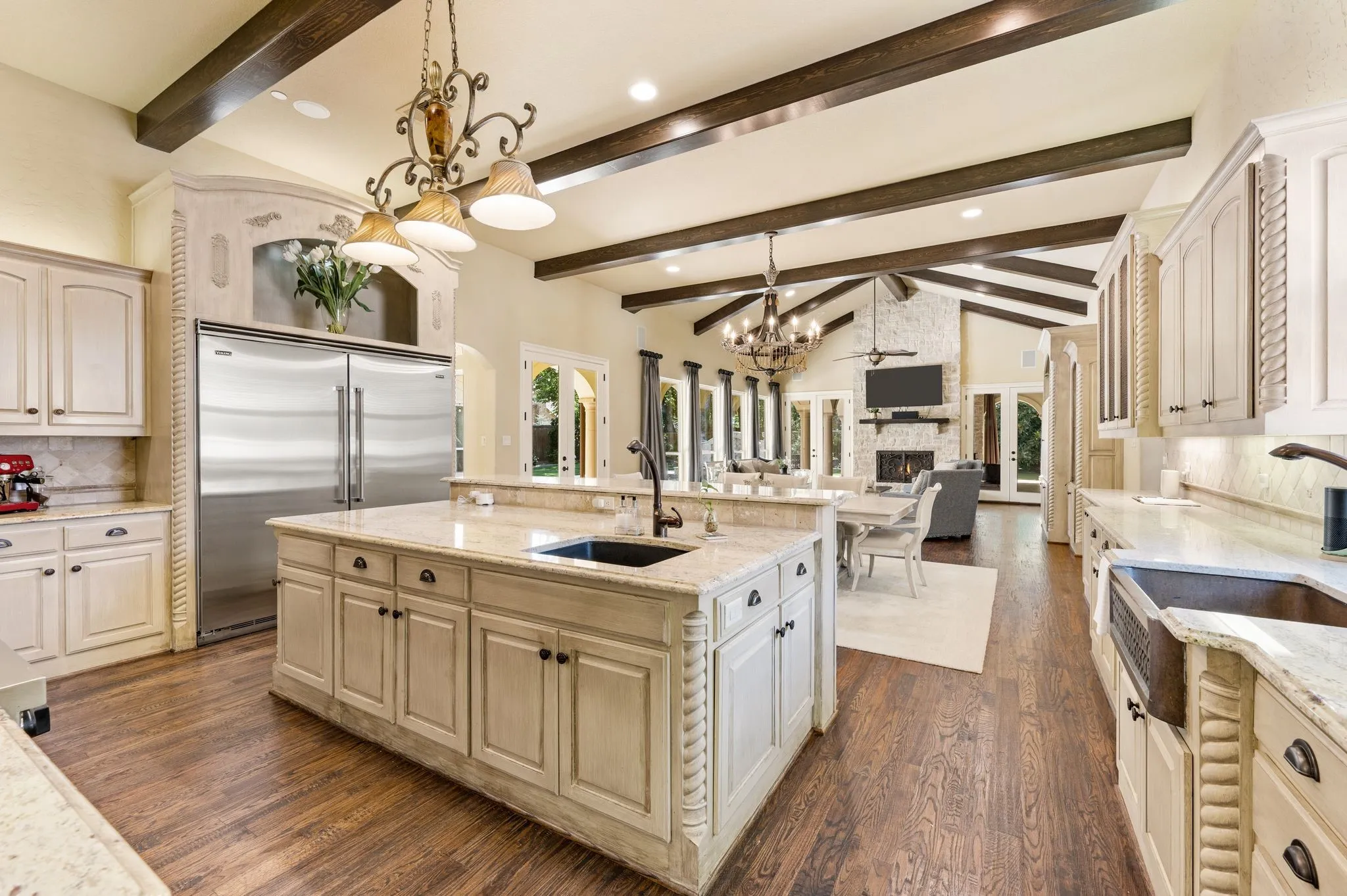 Spacious kitchen featuring an expansive center island with an undermount sink, built-in stainless steel refrigerator, white cabinetry, wood-finish flooring, and exposed ceiling beams