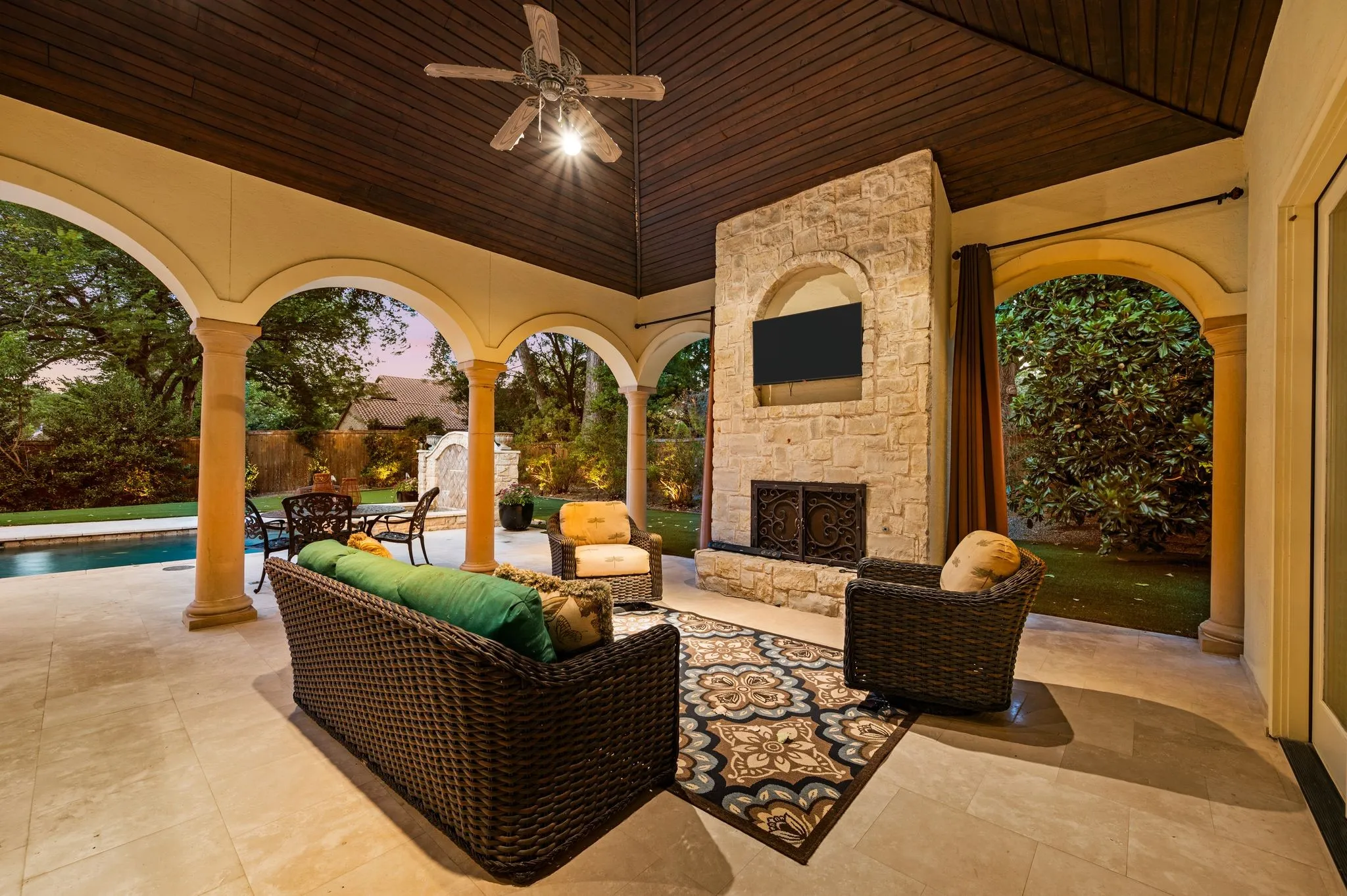 Covered patio with an arched colonnade, stone fireplace, wood-paneled ceiling, and a ceiling fan