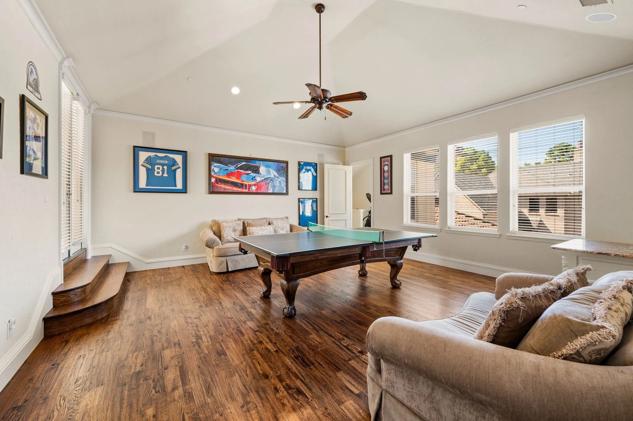 Expansive recreation room featuring rich wood-finish flooring, vaulted ceiling with ceiling fan, and abundant natural light from multiple large windows