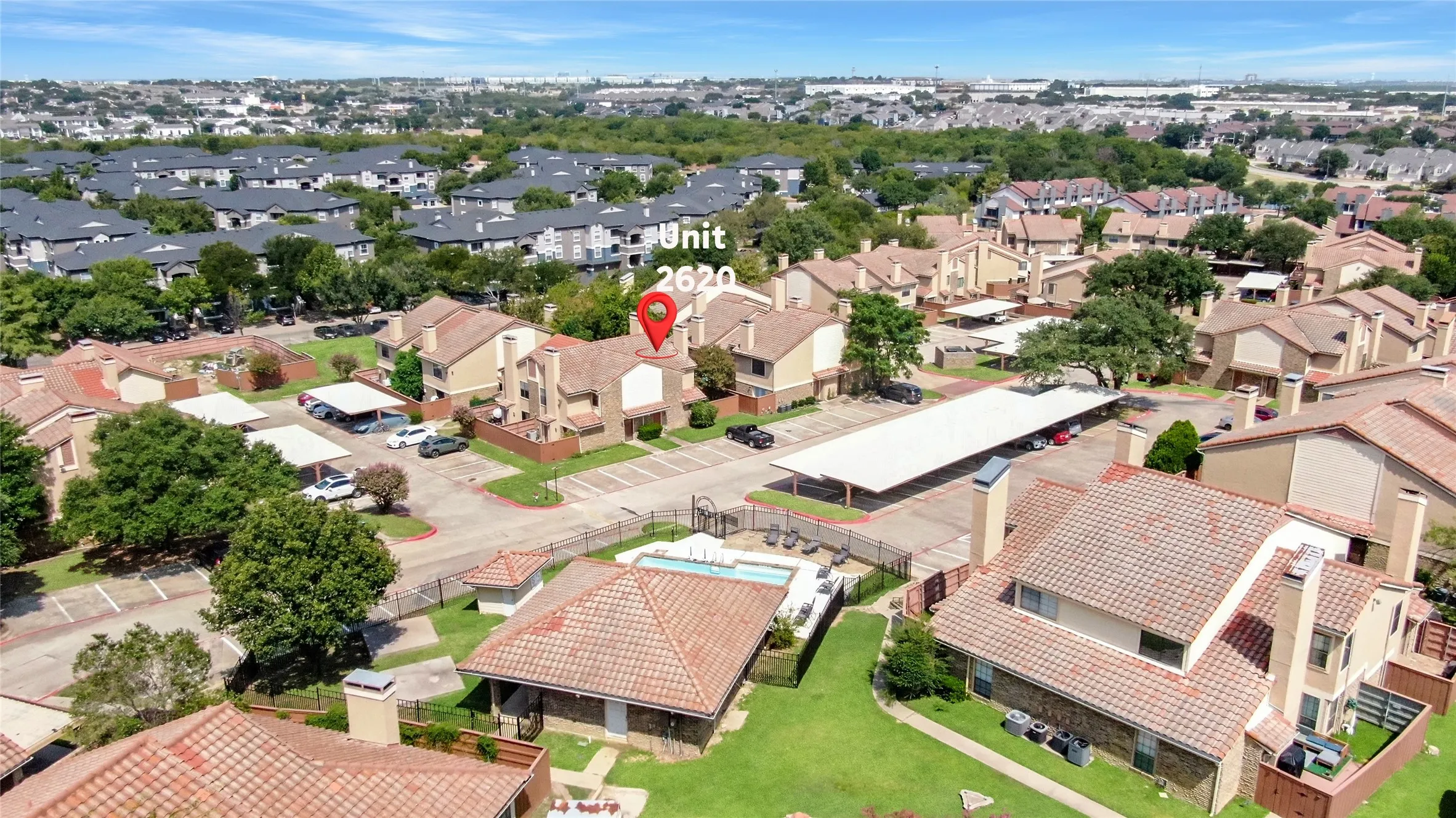 Community featuring multiple residential buildings with terracotta-style roofs, a central parking area with covered spaces, and a shared outdoor pool