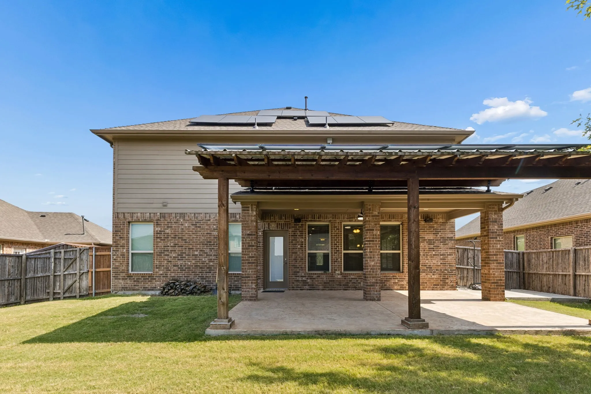 Expansive backyard featuring a covered patio with a wooden pergola, a brick and siding exterior, multiple windows, and rooftop solar panels