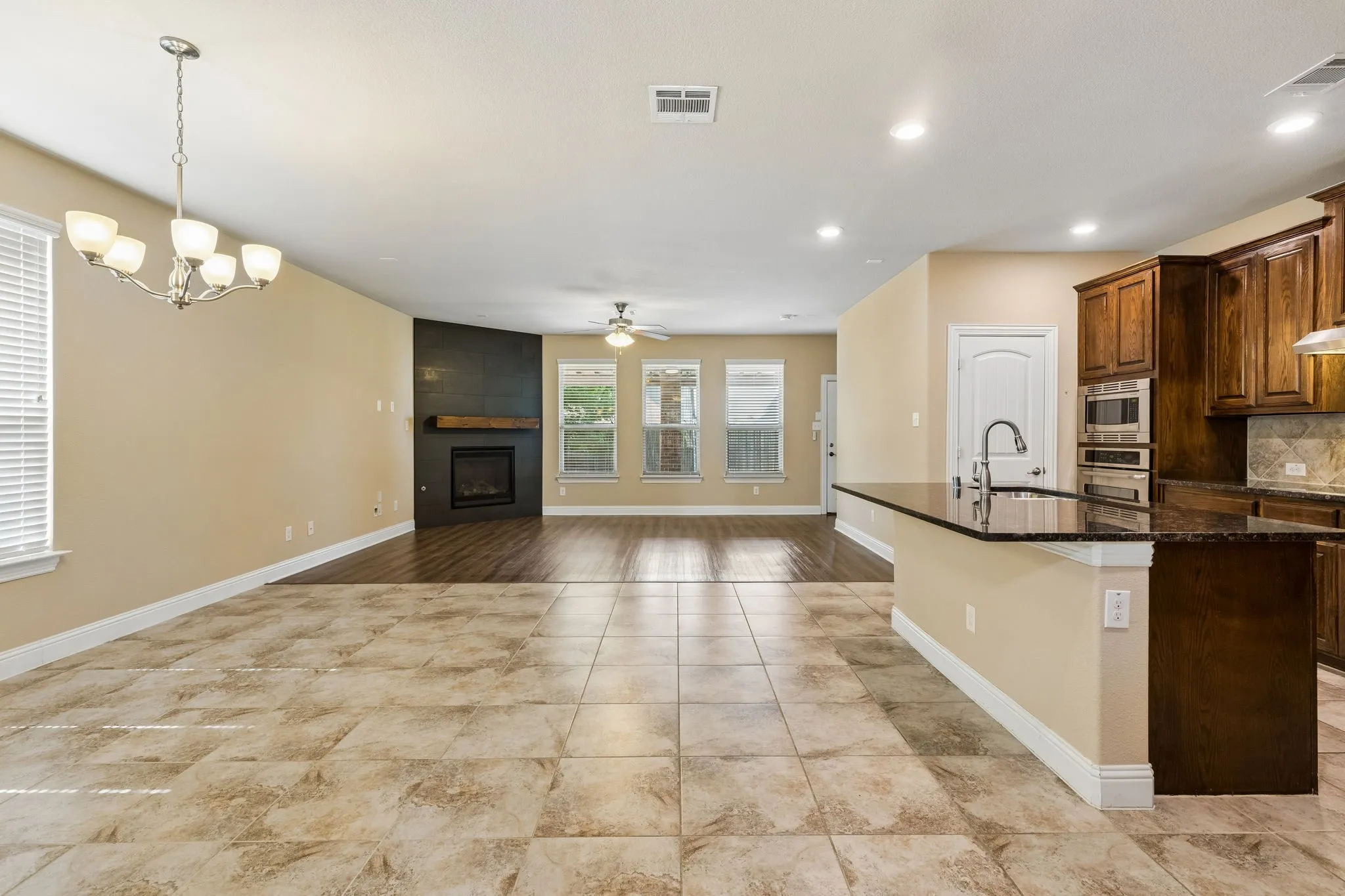 Spacious open-concept interior featuring light-toned tile flooring, a fireplace with a wood mantel, dark wood-finish cabinetry, built-in stainless steel appliances, and a kitchen island with a dark countertop