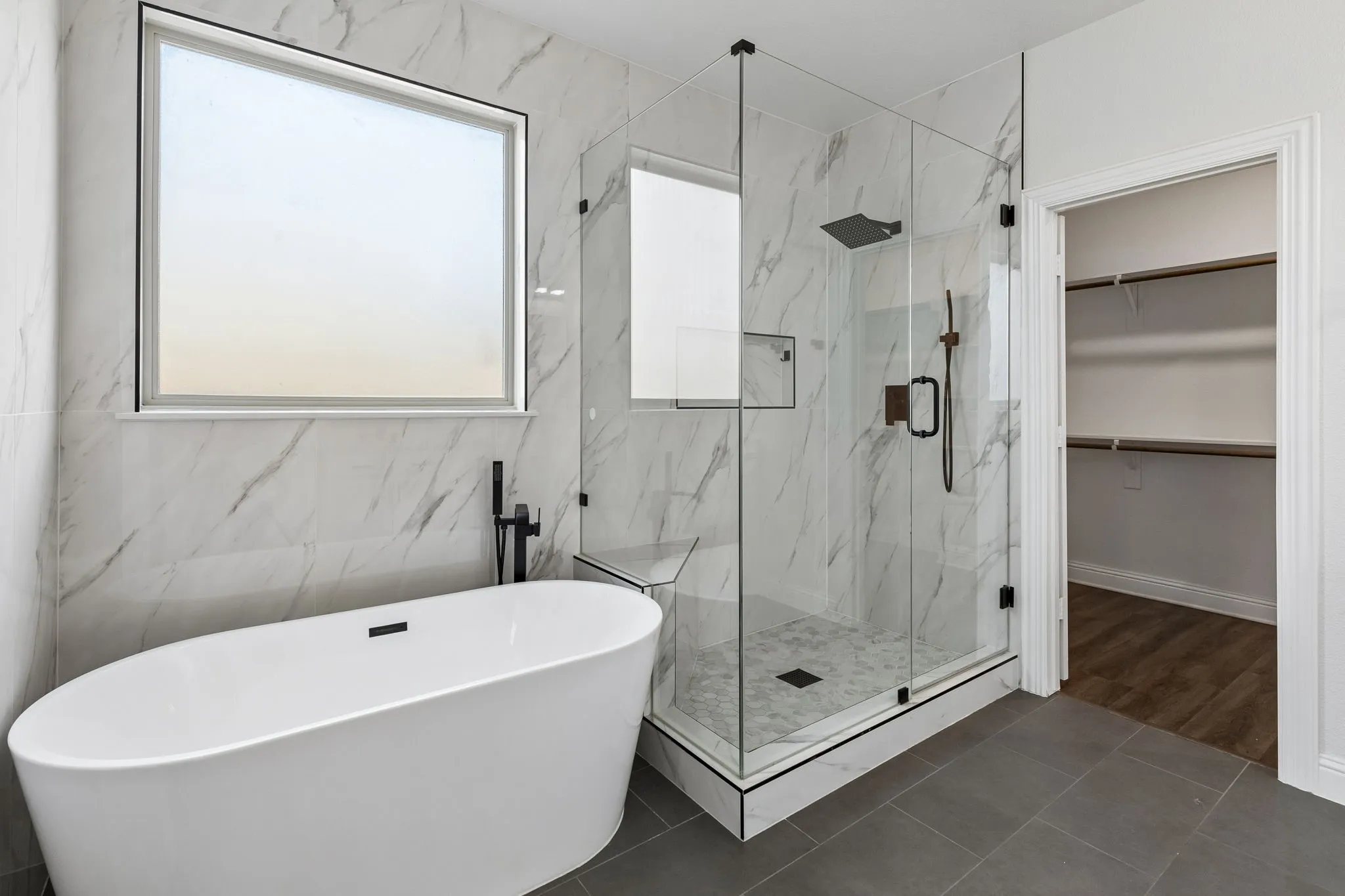 Freestanding soaking tub with a floor-mounted faucet, glass-enclosed shower with a built-in bench, wall-to-wall marble-patterned tile, and gray floor tile