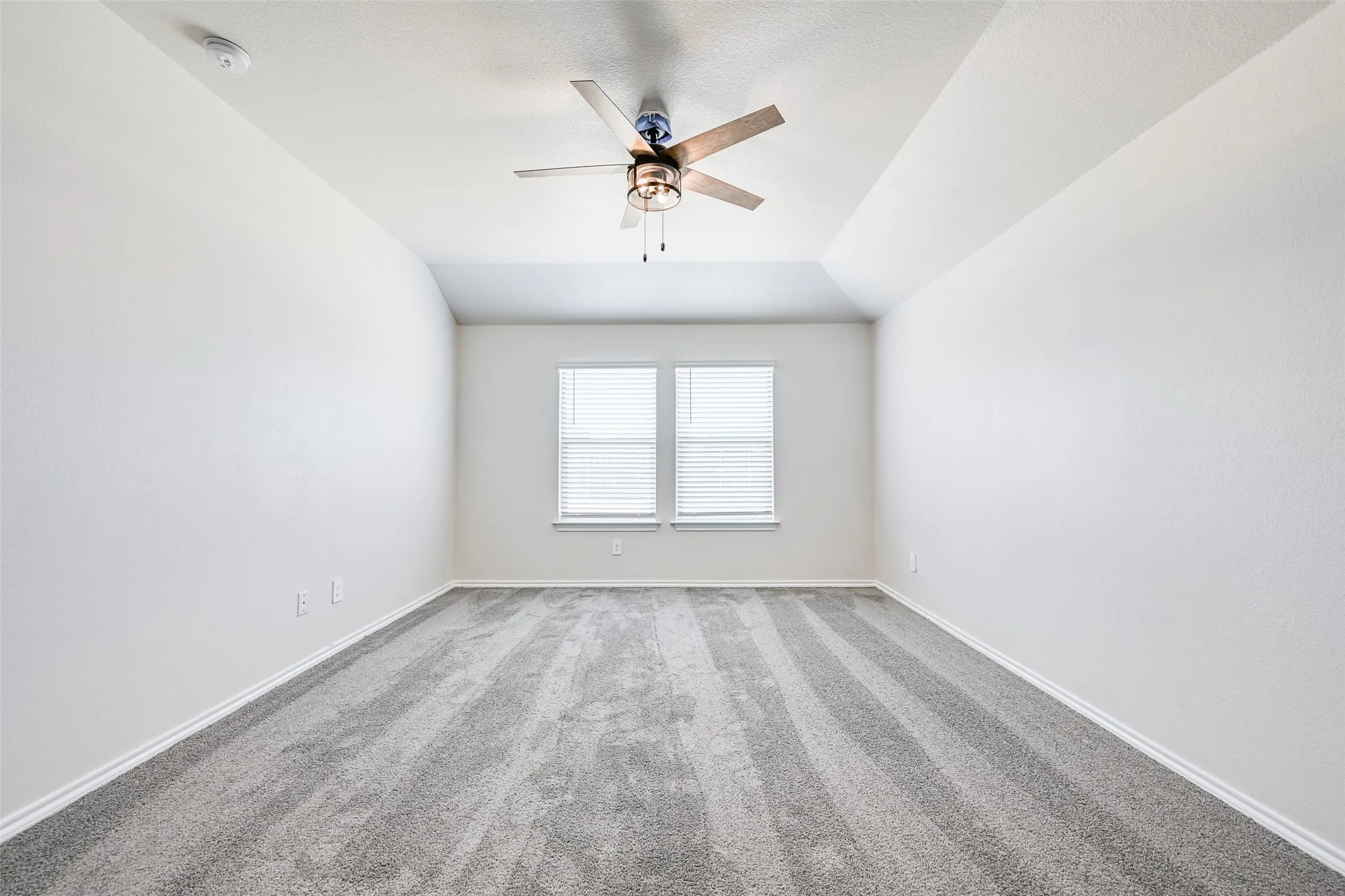 Spacious room featuring vaulted ceilings, a ceiling fan with integrated lighting, twin windows with blinds, and light-toned carpeting