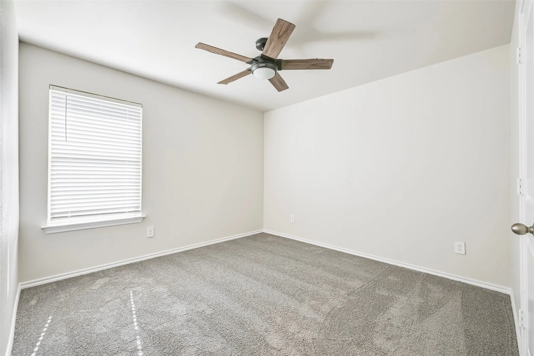 Recessed window with white blinds, grey carpeting, white painted walls, and ceiling fan with wood-finish blades