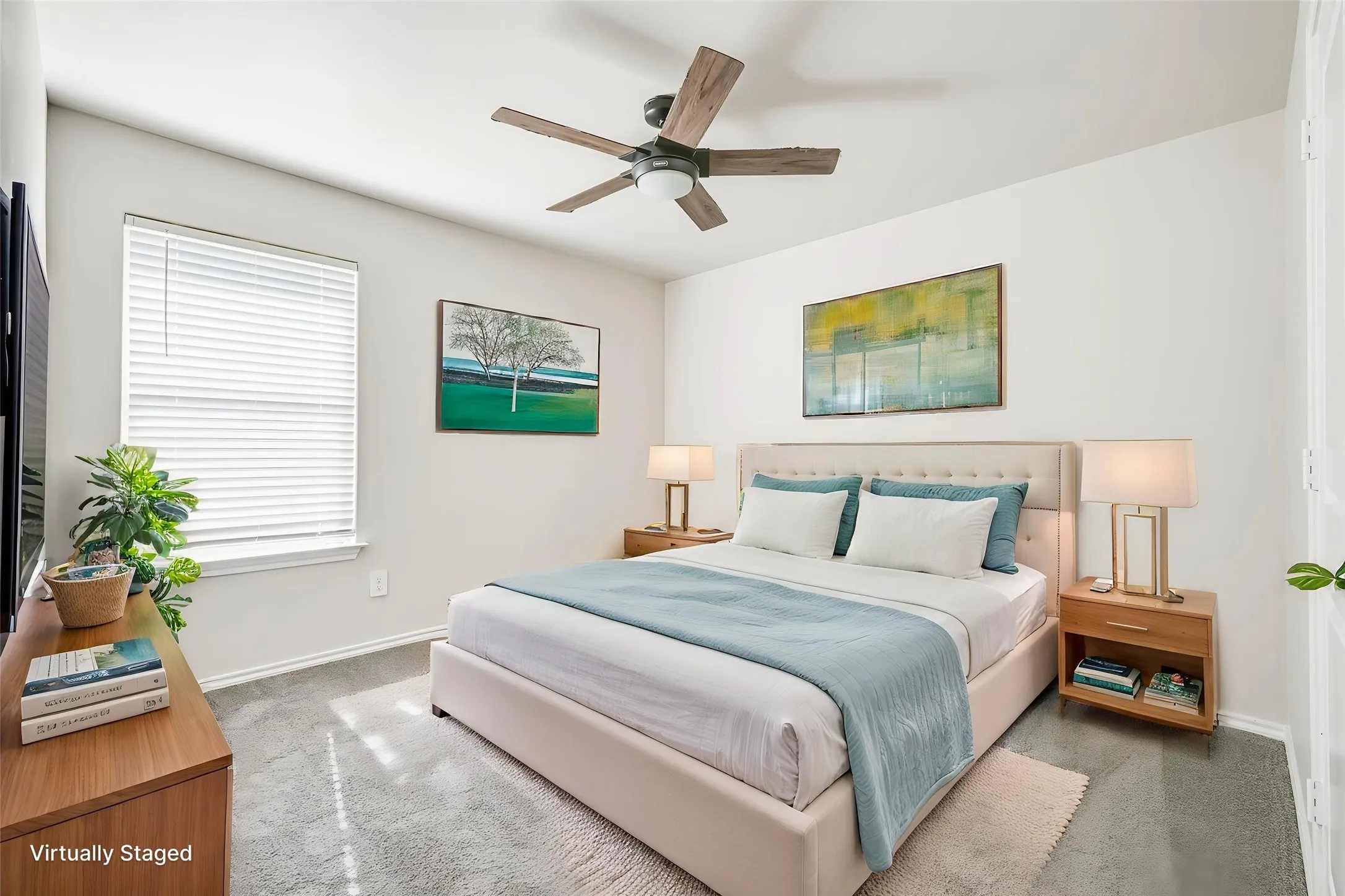 Bright primary bedroom featuring a ceiling fan, large windows with plantation shutters, wood-finish nightstands with integrated shelving, neutral wall-to-wall carpeting, and artwork accents
