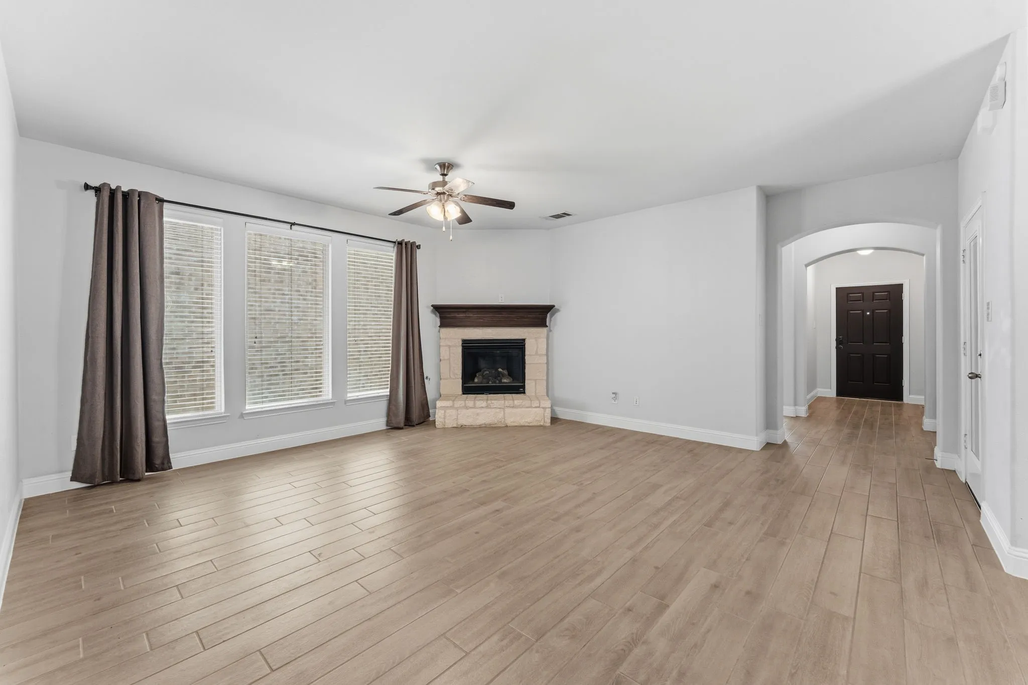 Spacious living area featuring wood-finish flooring, a corner fireplace with stone surround and wood mantel, multiple windows, and an arched entry to the foyer