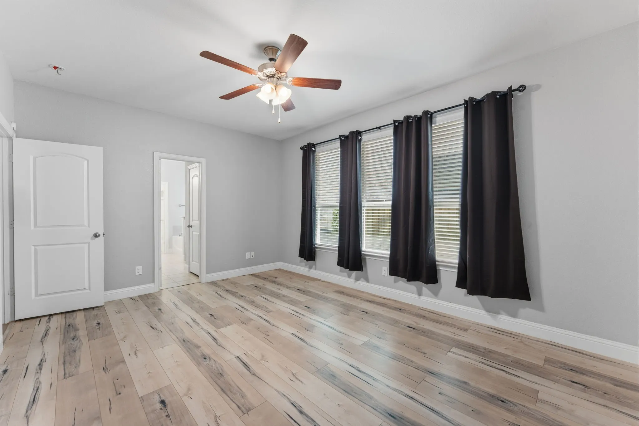 Spacious room featuring light wood-finish flooring, a ceiling fan with integrated lighting, and three windows with blinds