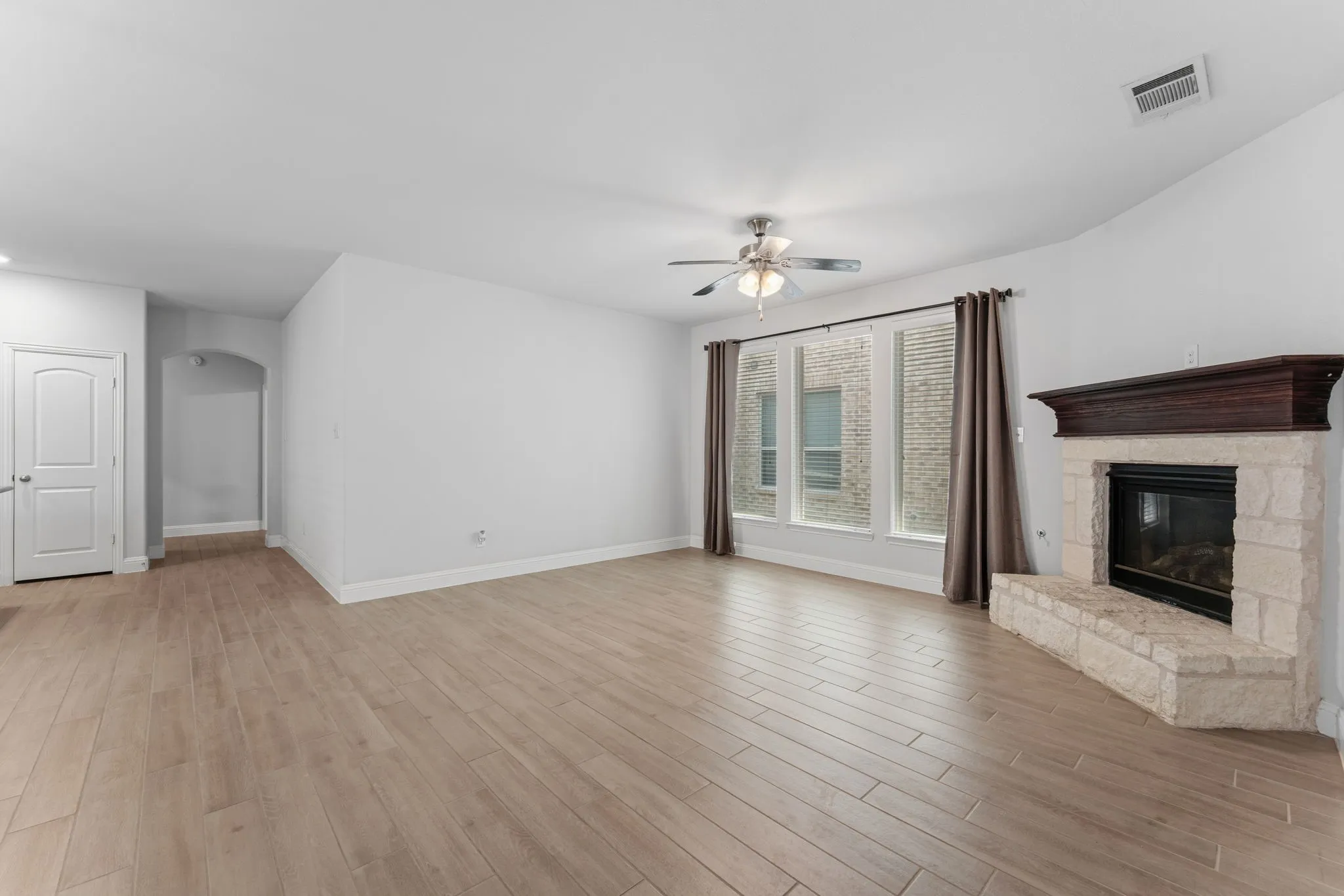 Spacious living area featuring wood-finish flooring, a stone fireplace with a dark wood mantel, and a bank of three large windows