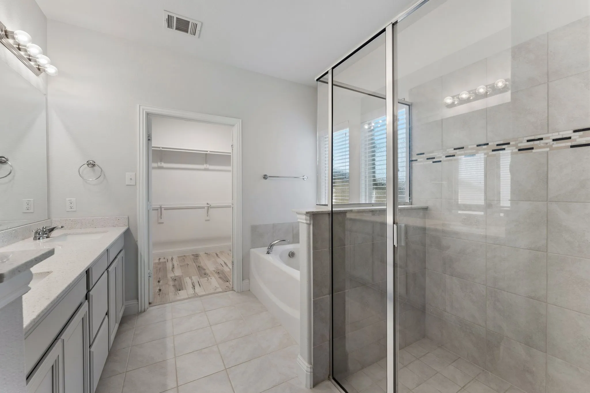 Modern bathroom featuring a dual vanity with light-toned cabinetry and countertops, a built-in soaking tub, a glass-enclosed shower with tiled walls and decorative accent band, and an adjacent walk-in closet with wood-finish flooring and shelving