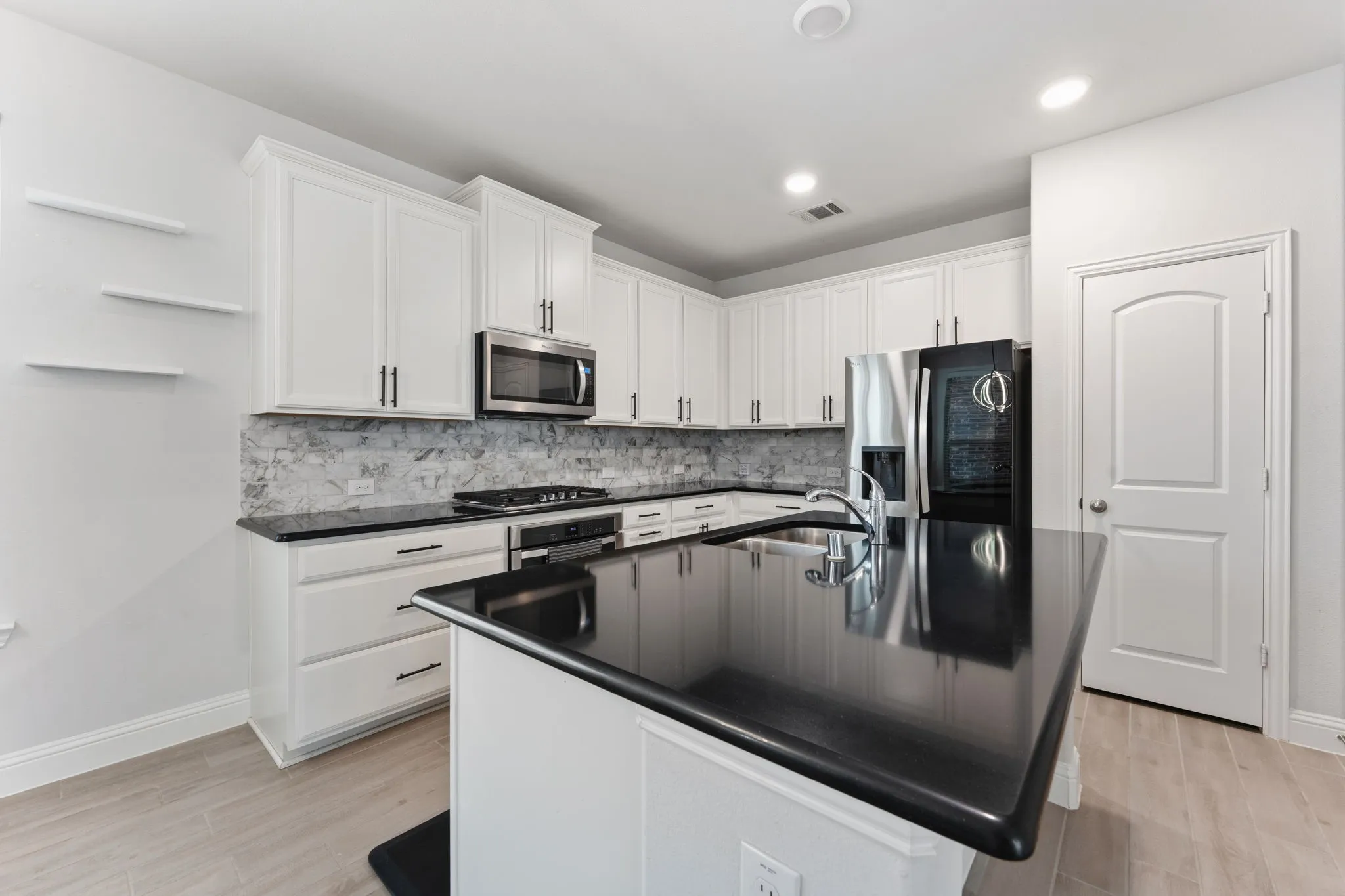 Kitchen featuring an island with an undermount sink, black countertops, stainless steel appliances, white cabinetry, and a patterned tile backsplash