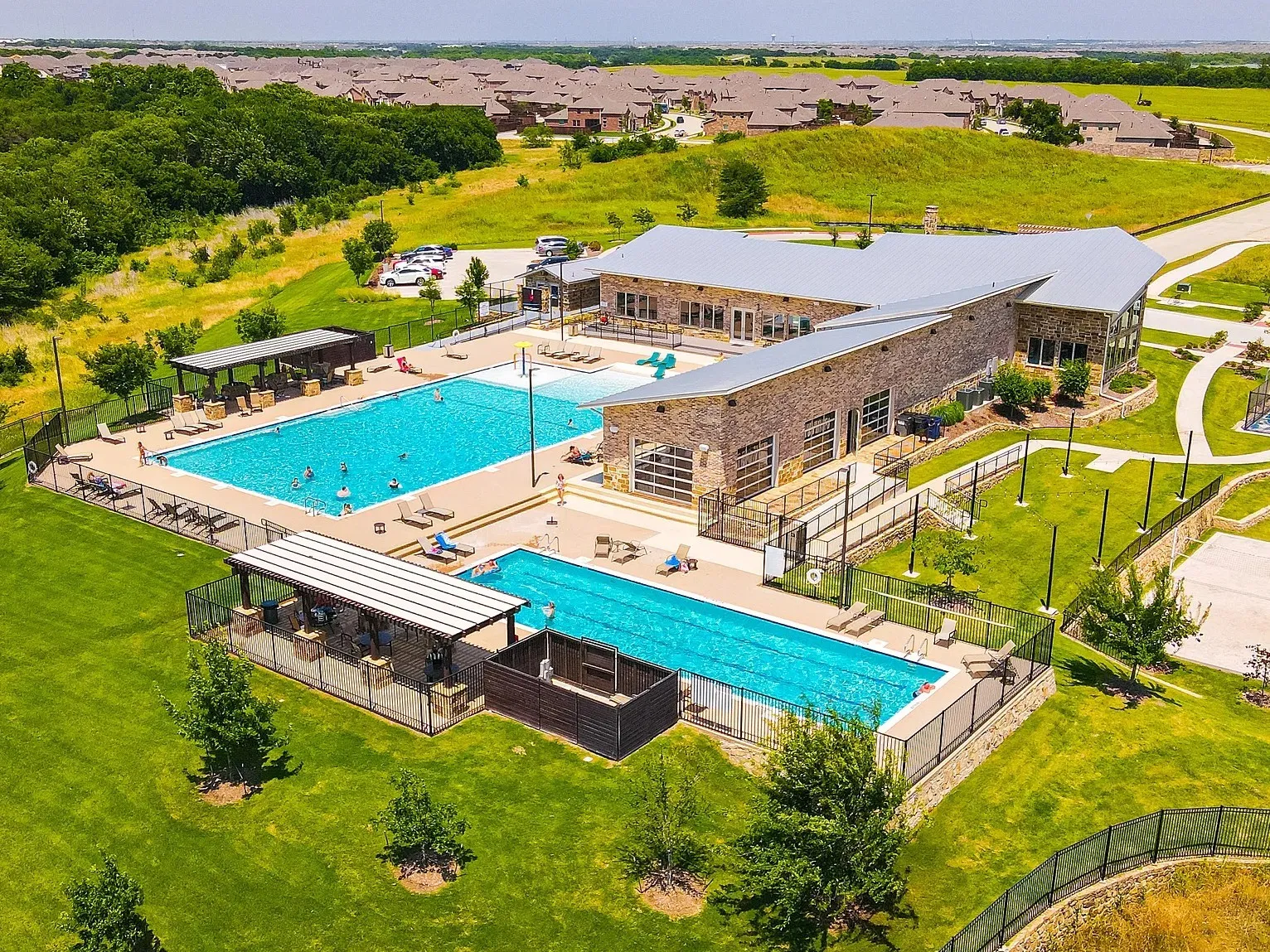 Community clubhouse with a stone exterior, multiple outdoor swimming pools, shaded cabana areas, and expansive green spaces