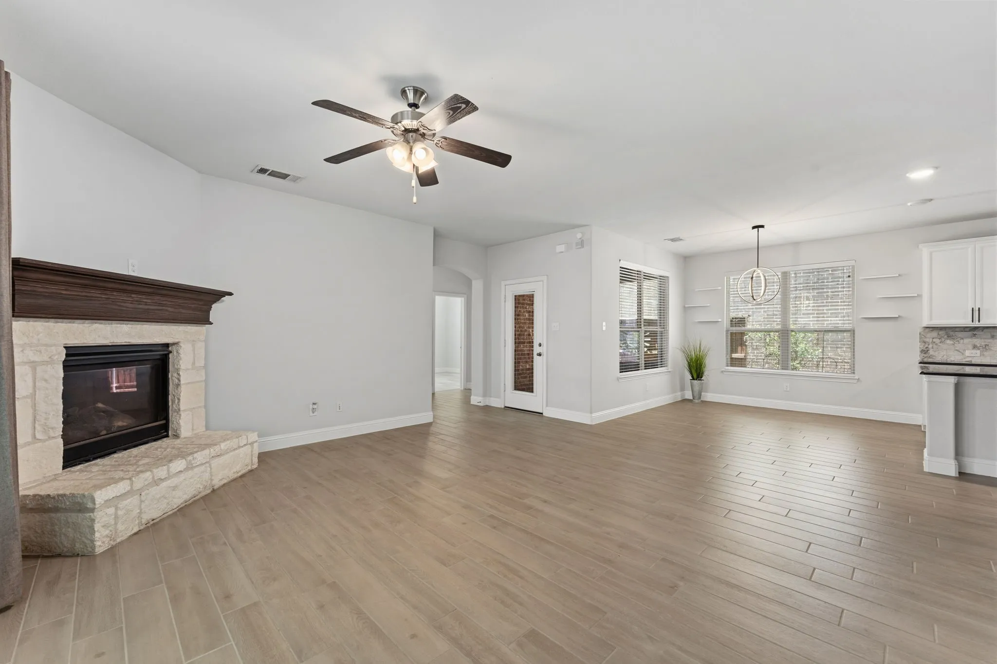 Spacious living area featuring wood-finish flooring, a stone fireplace with a dark wood mantel, and a ceiling fan with integrated lighting
