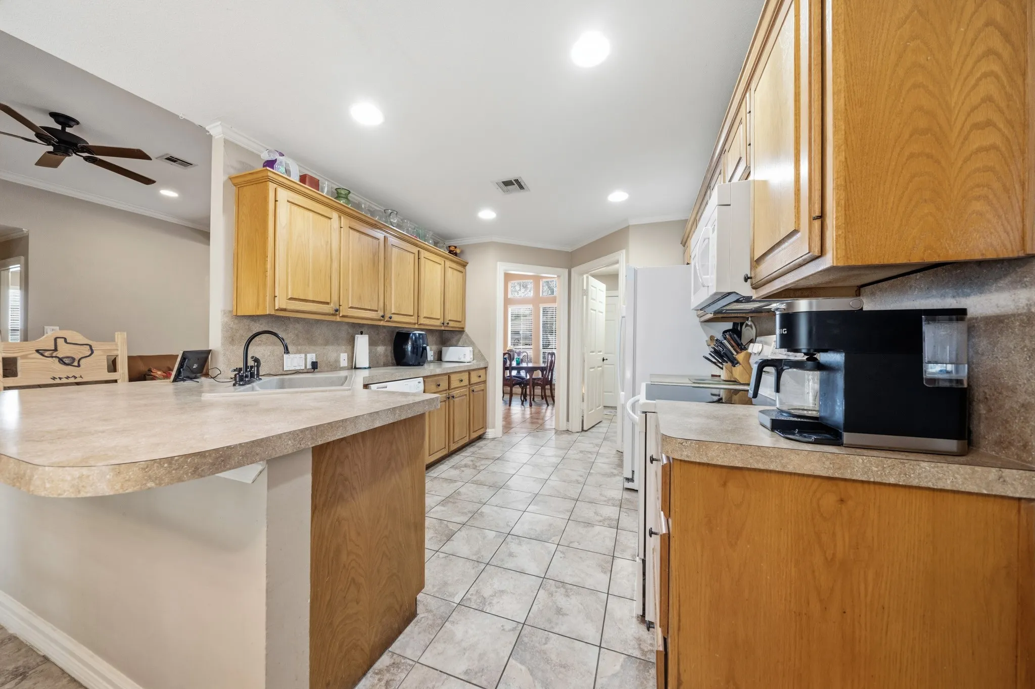 Spacious kitchen featuring wood-finish cabinetry, light stone countertops with curved island seating, recessed ceiling lighting, tile flooring, and stainless steel appliances