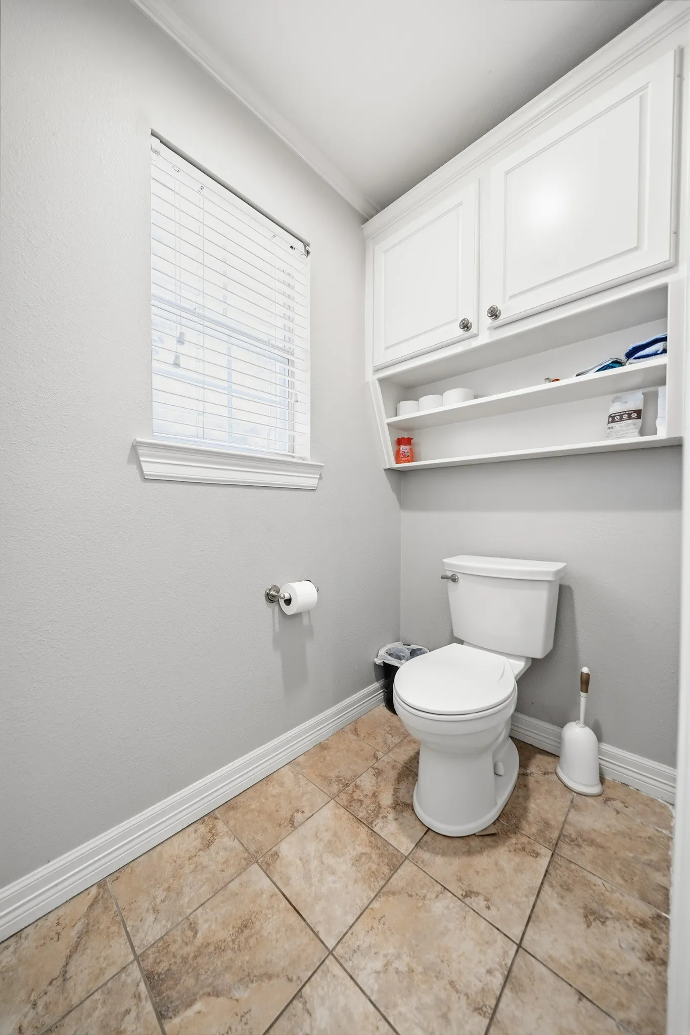 Compact bathroom featuring ceramic tile flooring, white painted cabinetry with shelving, a one-piece toilet with tank, chrome towel bar, and a horizontal blinds window for natural light