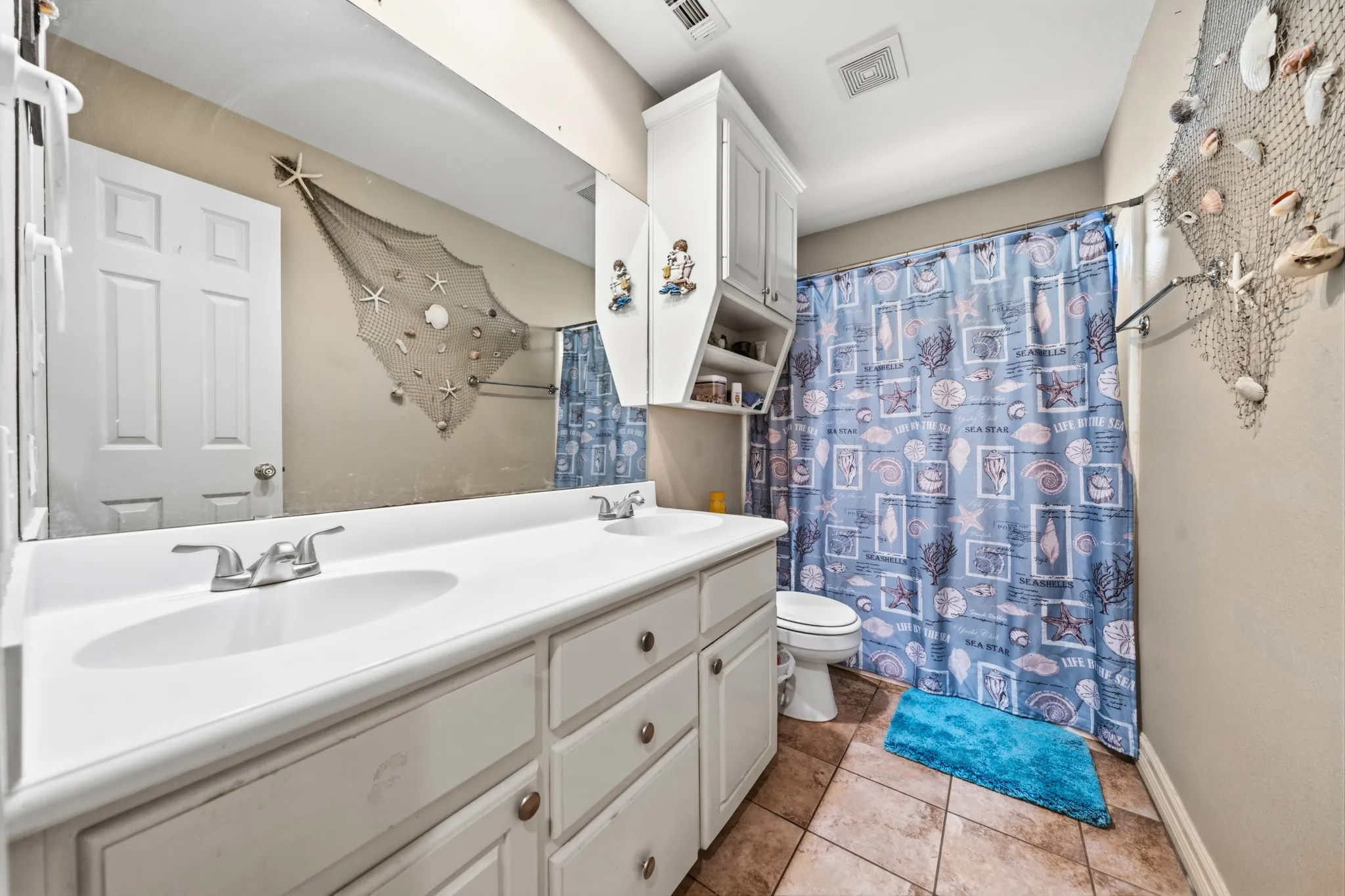 Spacious bathroom featuring a long vanity with dual sinks, ample countertop storage with cabinetry, ceramic tile flooring, and a shower-tub combination with blue tile surround and coordinating shower curtain