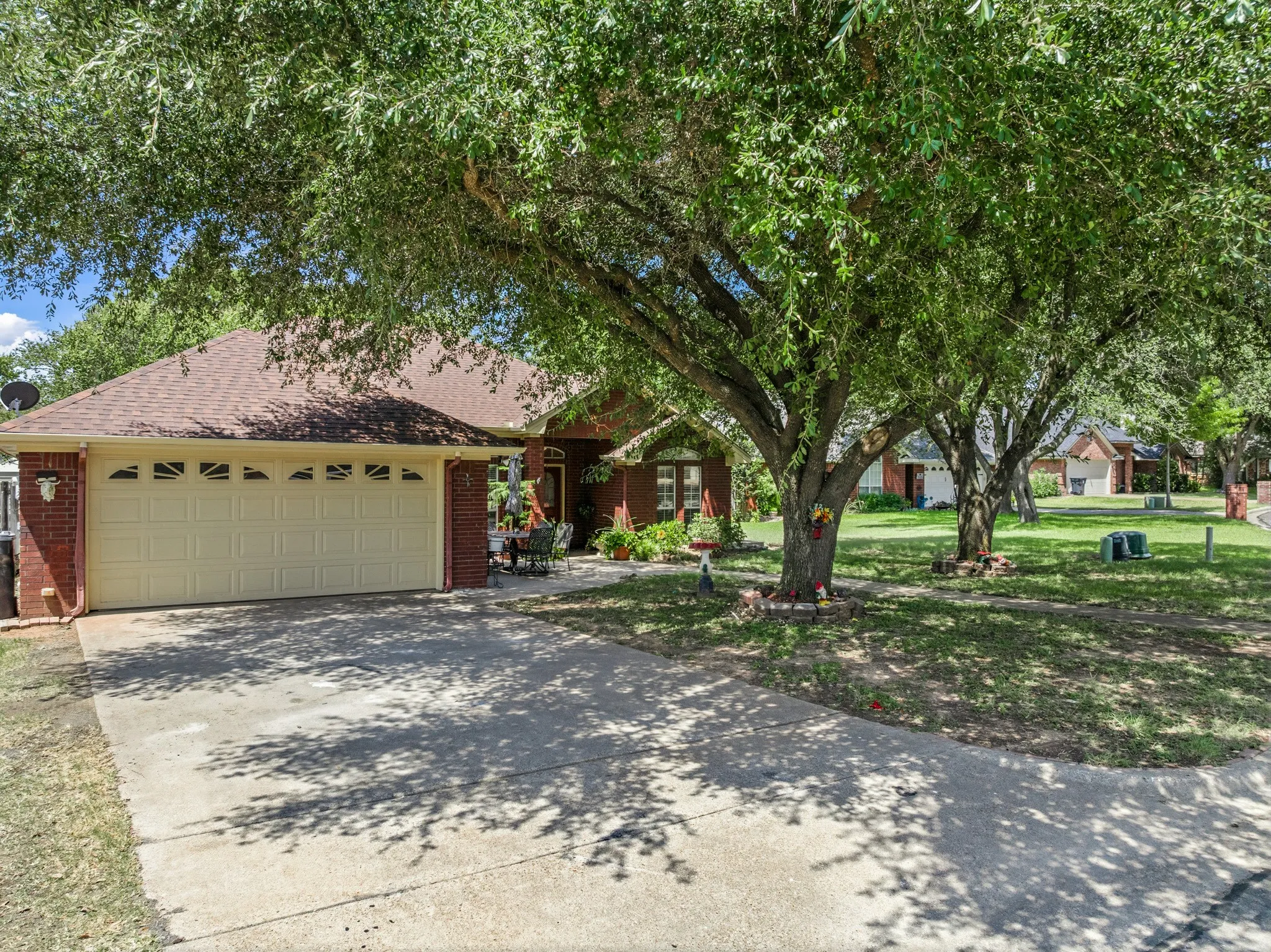 Red brick exterior with a tan garage door, brown shingle roof, paved driveway, and mature trees
