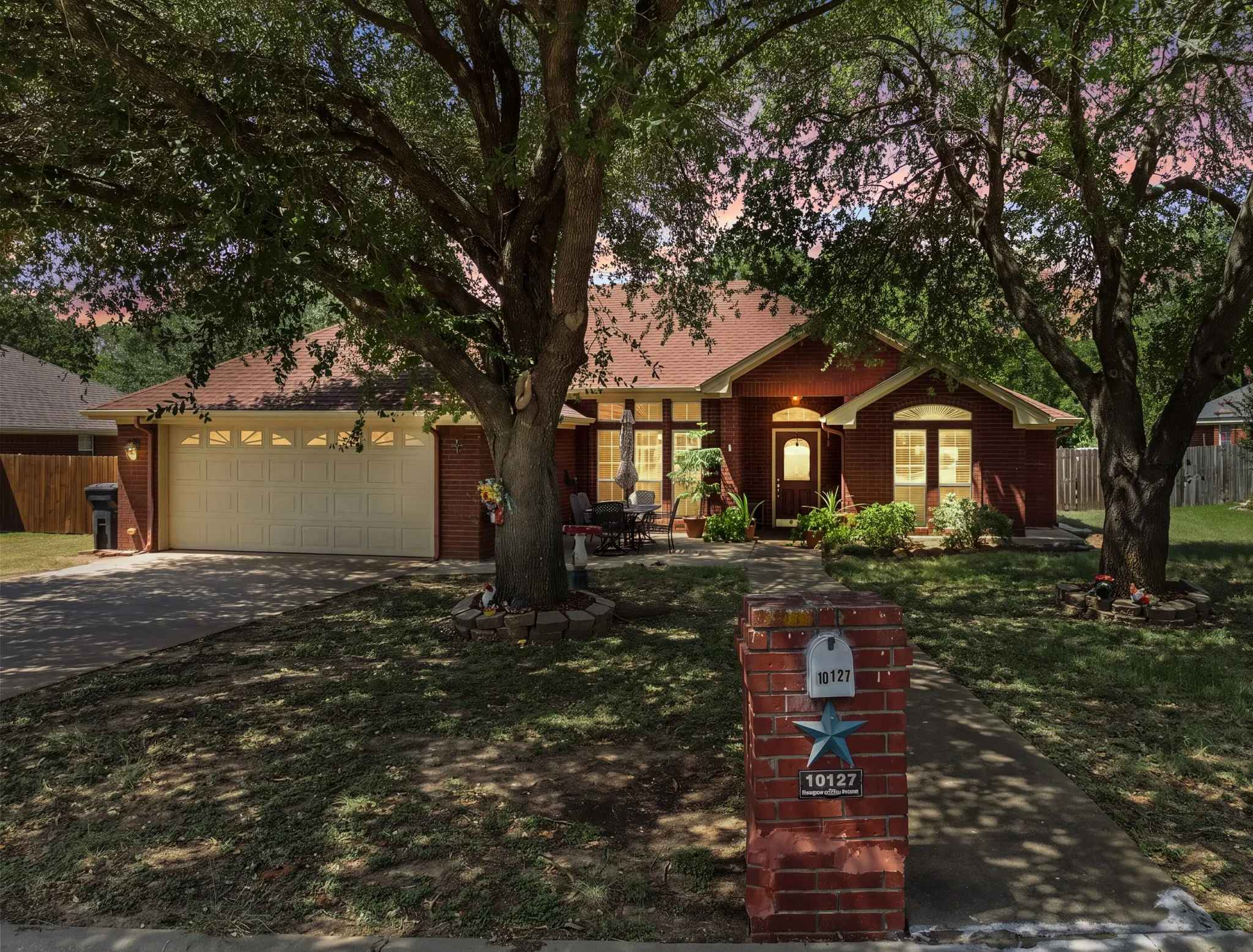 Charming brick residence nestled beneath mature shade trees, featuring a welcoming front elevation with gabled rooflines, large windows with natural light, and a two-car garage