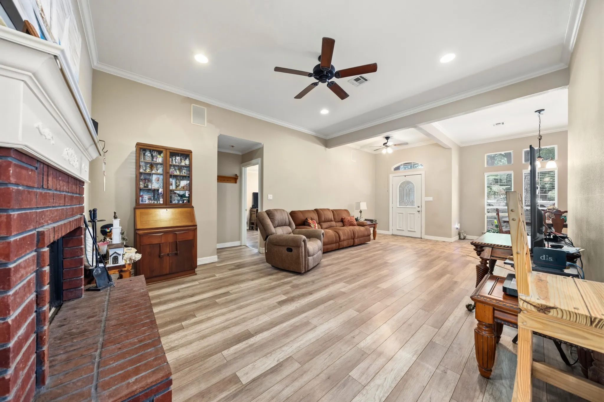 Spacious open-concept living area featuring wood-finish plank flooring, coffered ceiling with ceiling fan, recessed lighting, and a wood-burning fireplace with brick surround