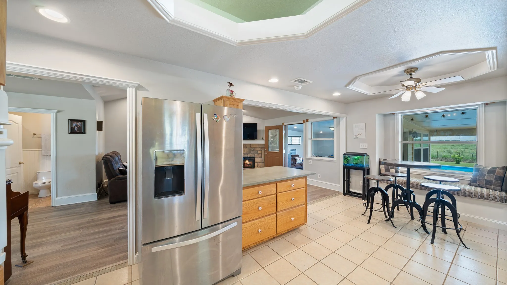 Open-concept kitchen and dining area featuring stainless steel side-by-side refrigerator, wood-finish cabinetry with ample storage, tile countertops and flooring, ceiling fan, and large windows framing scenic outdoor vistas